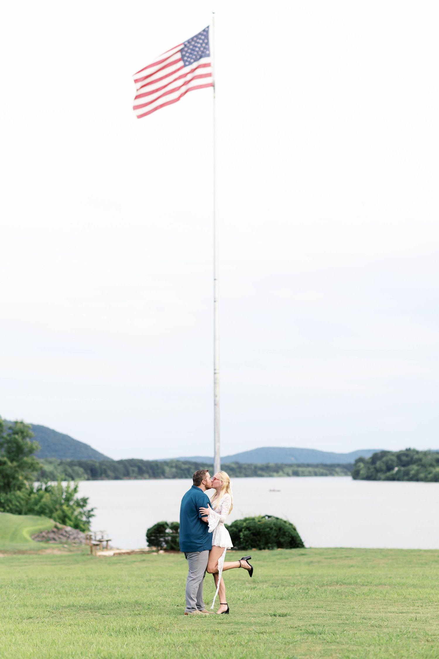 A couple kisses beneath a tall American flag pole on a green lawn beside a scenic river.