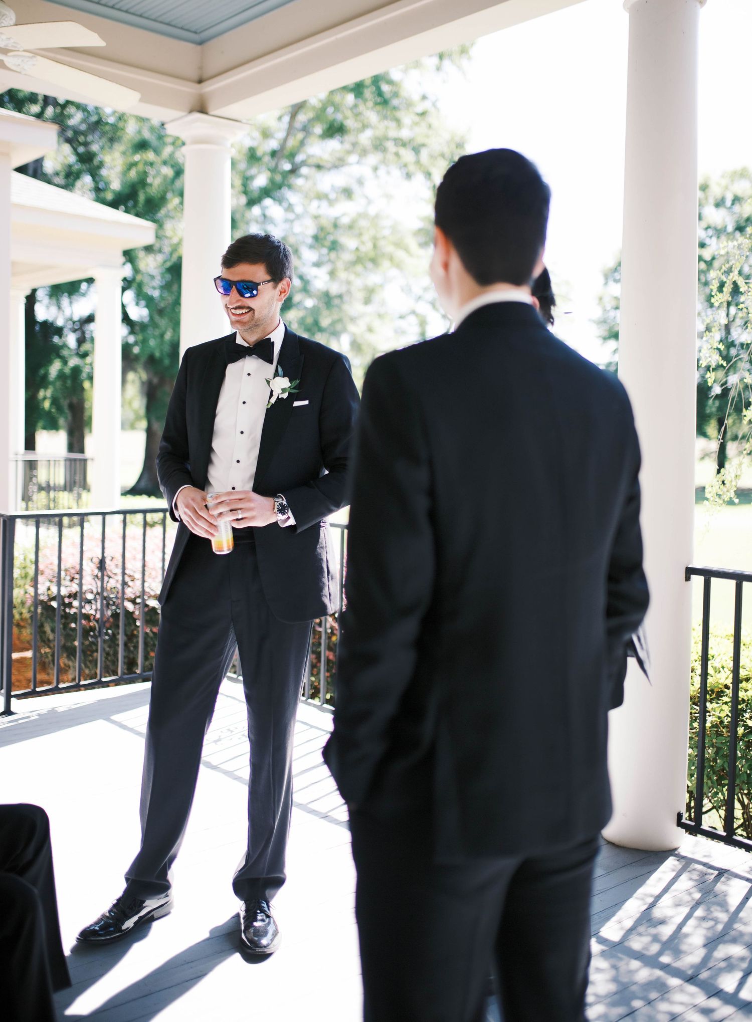 Two groomsmen in black tuxedos stand on white-columned porch during outdoor wedding celebration.