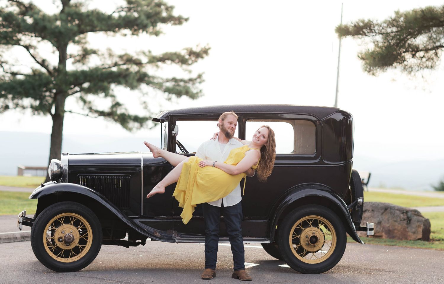 Couple posing with a vintage black Model T Ford car, man holding woman in yellow dress, outdoor scenic background.