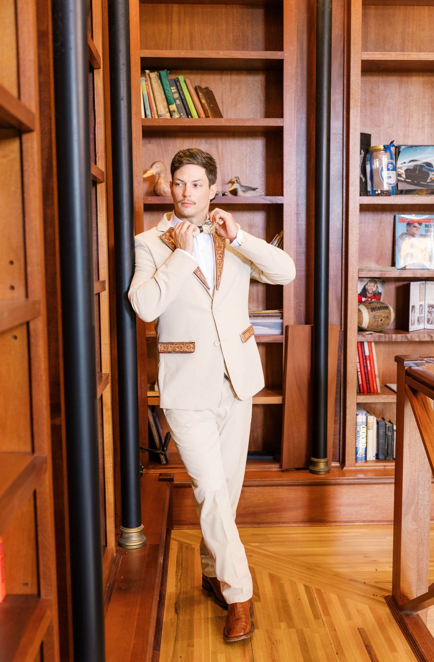 Man in cream suit adjusting bow tie in wood-paneled library with bookshelves.