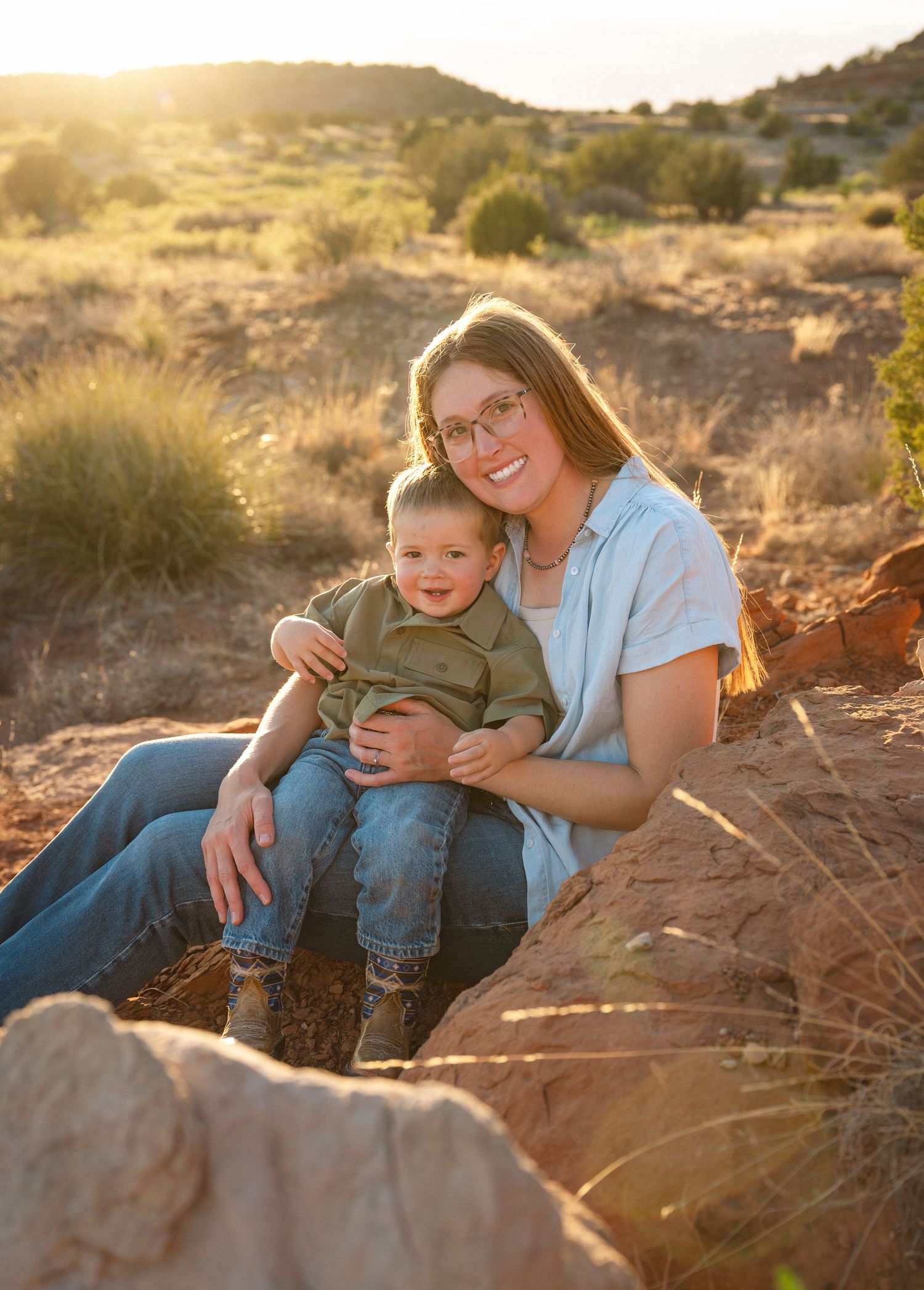 A smiling woman with glasses holds a toddler boy on red rocks in a sunny desert landscape at golden hour.