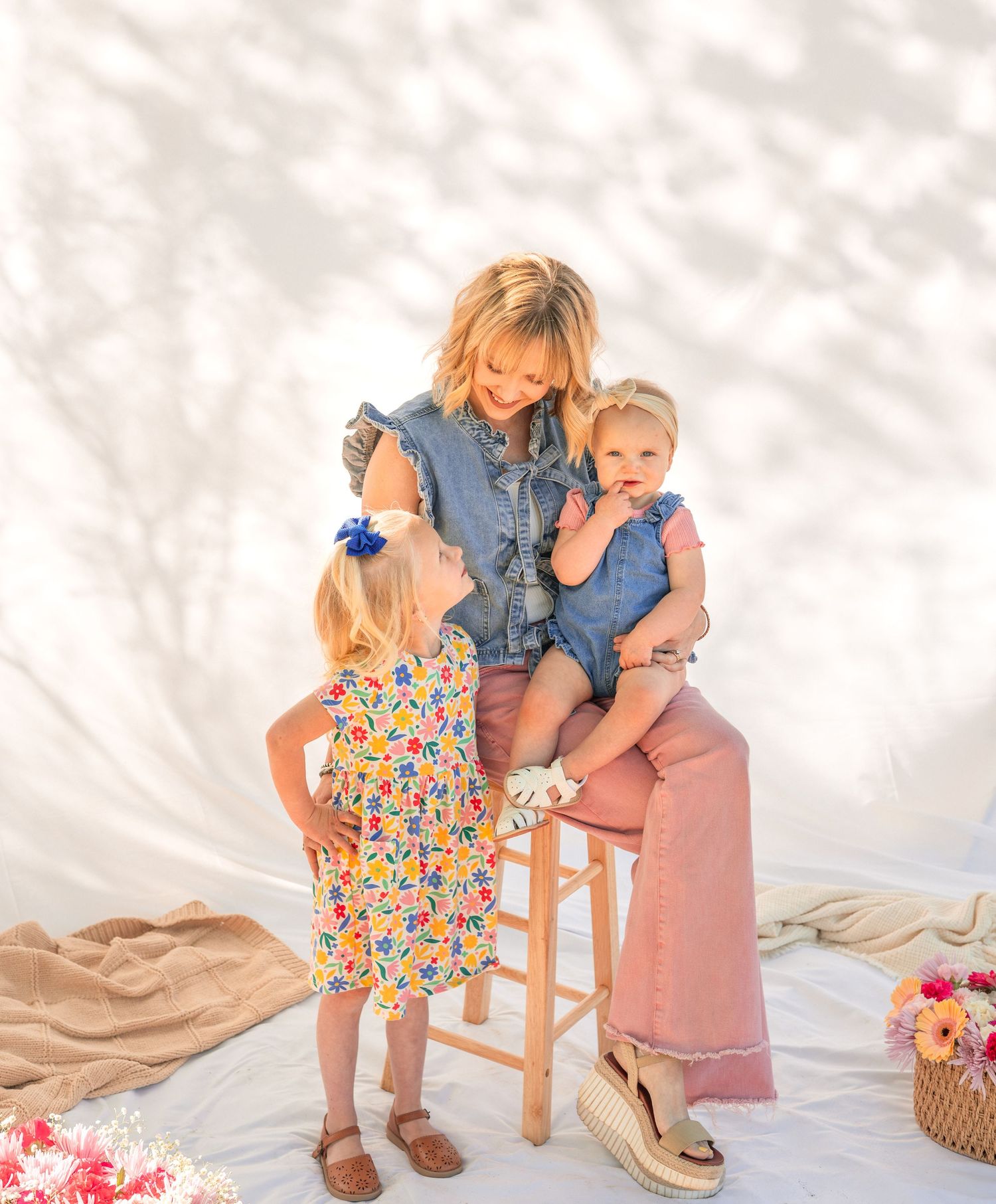 A mother in denim sits on a stool holding a baby while a young girl in floral dress stands nearby.