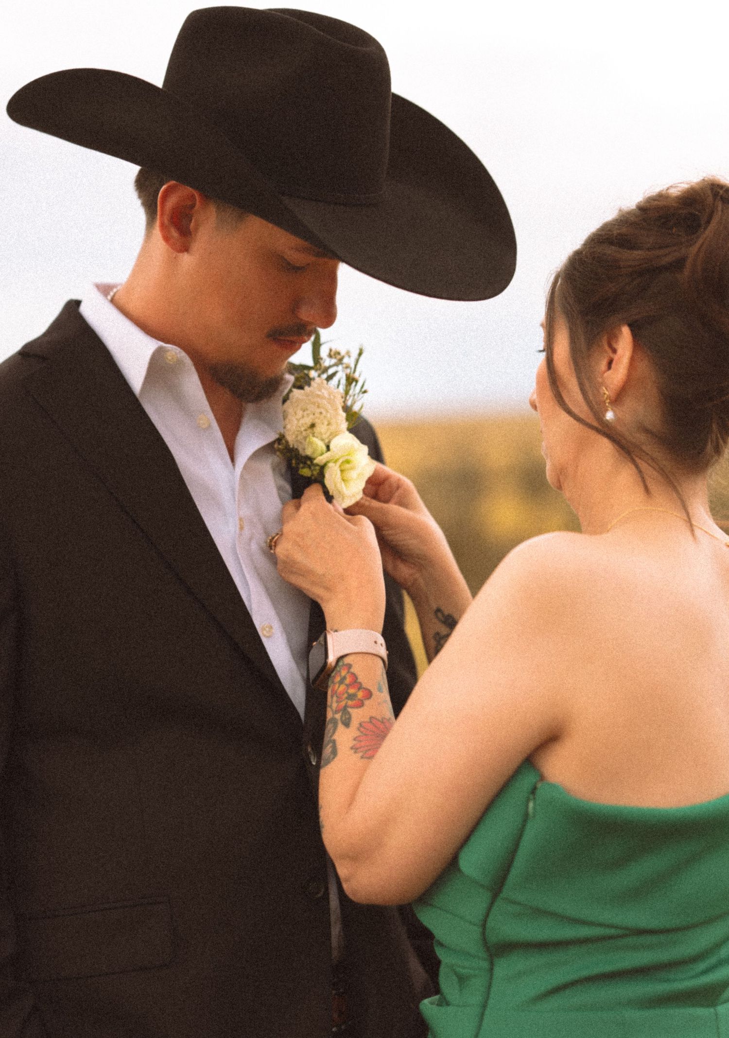 Woman with tattooed arm in green dress pinning boutonniere on groom in black cowboy hat and suit.