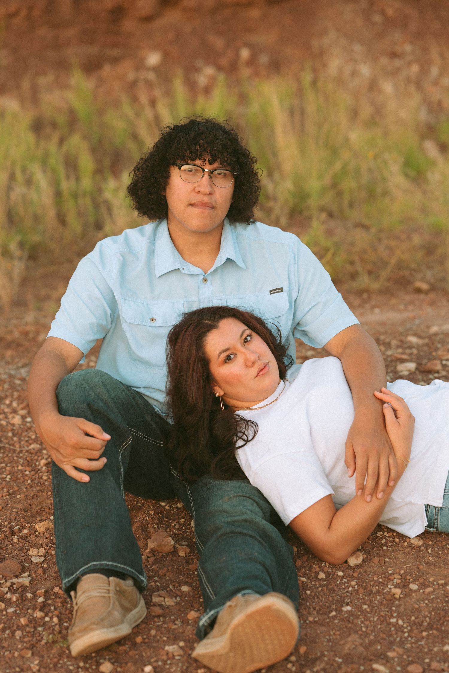 Couple sitting on red dirt ground, woman leaning against man, with green grass backdrop.