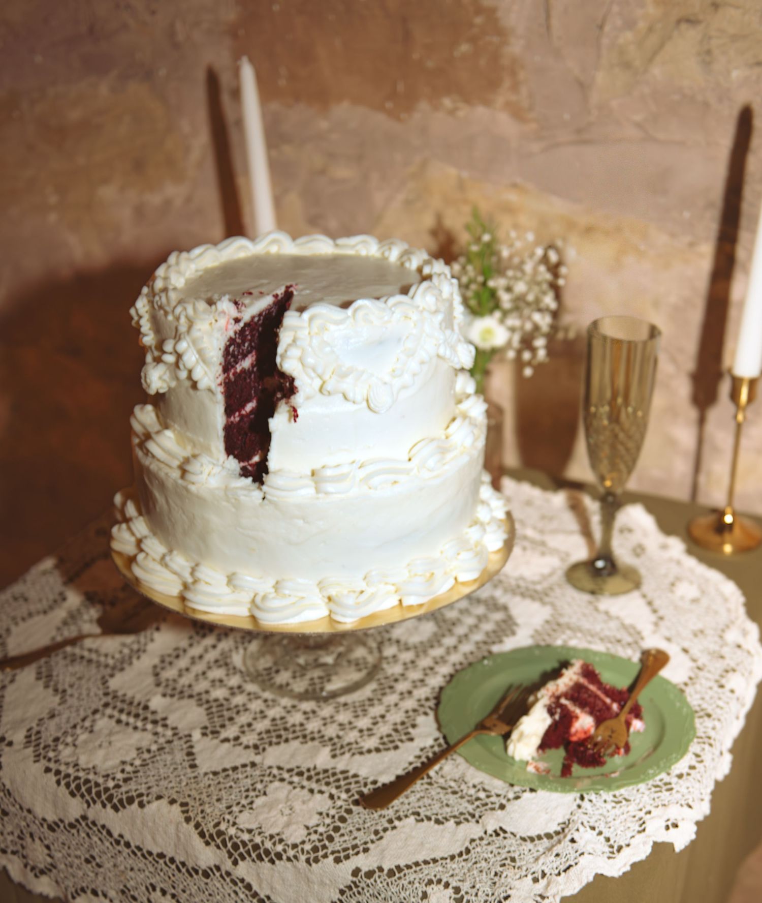 Two-tier white frosted red velvet wedding cake with floral decor and champagne flutes on lace tablecloth.