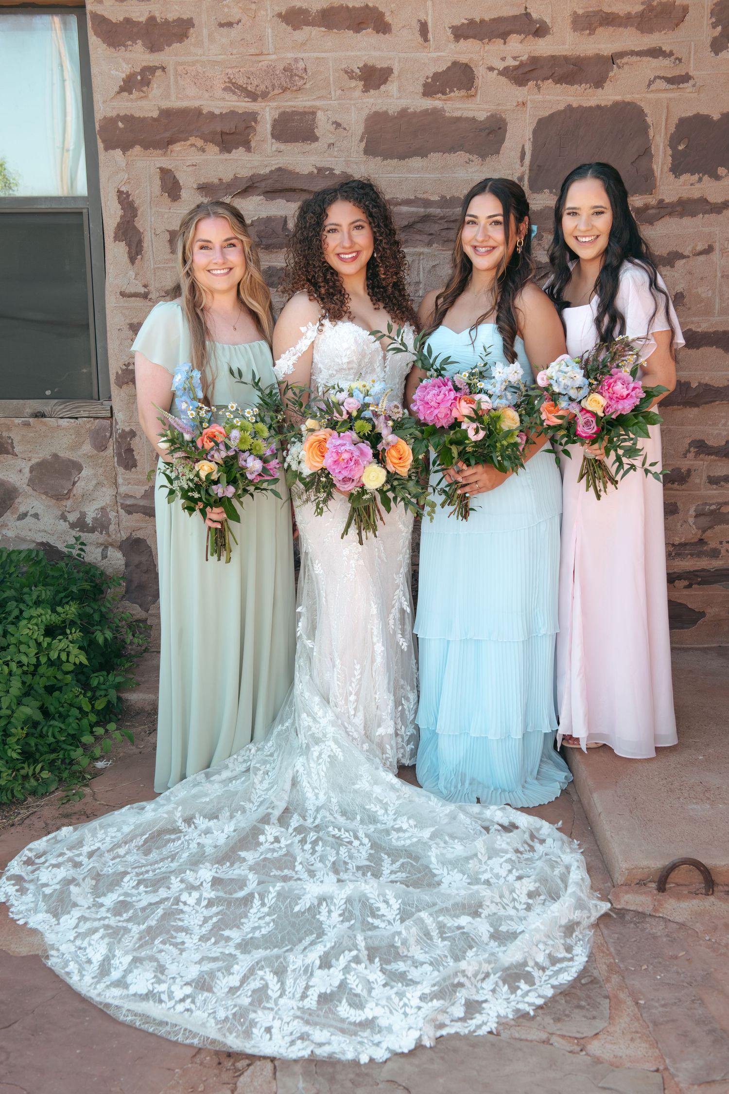 A bride in a lace gown poses with three bridesmaids in pastel dresses holding colorful bouquets.