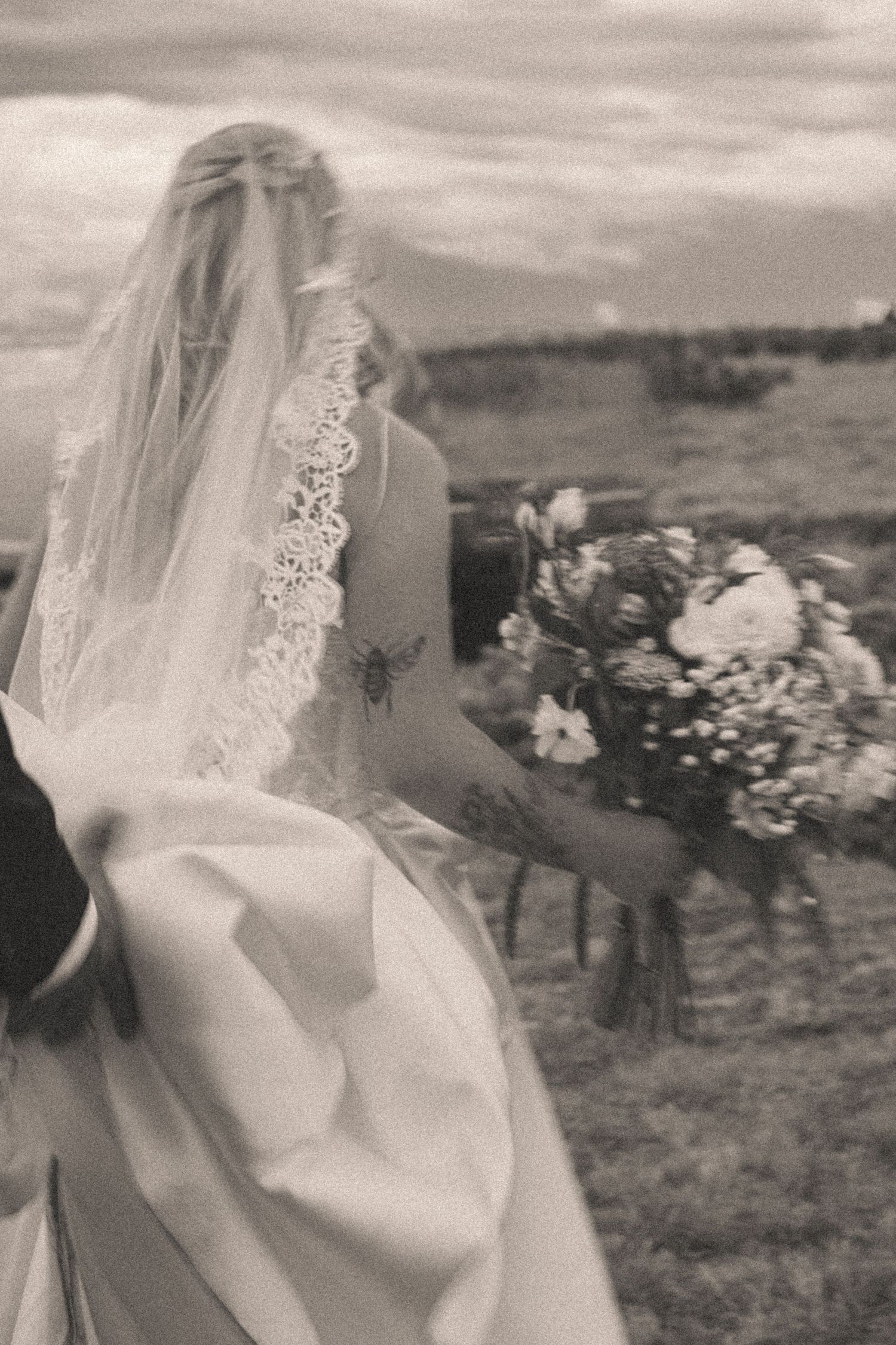 Black and white photo of bride in lace veil holding floral bouquet in open field.