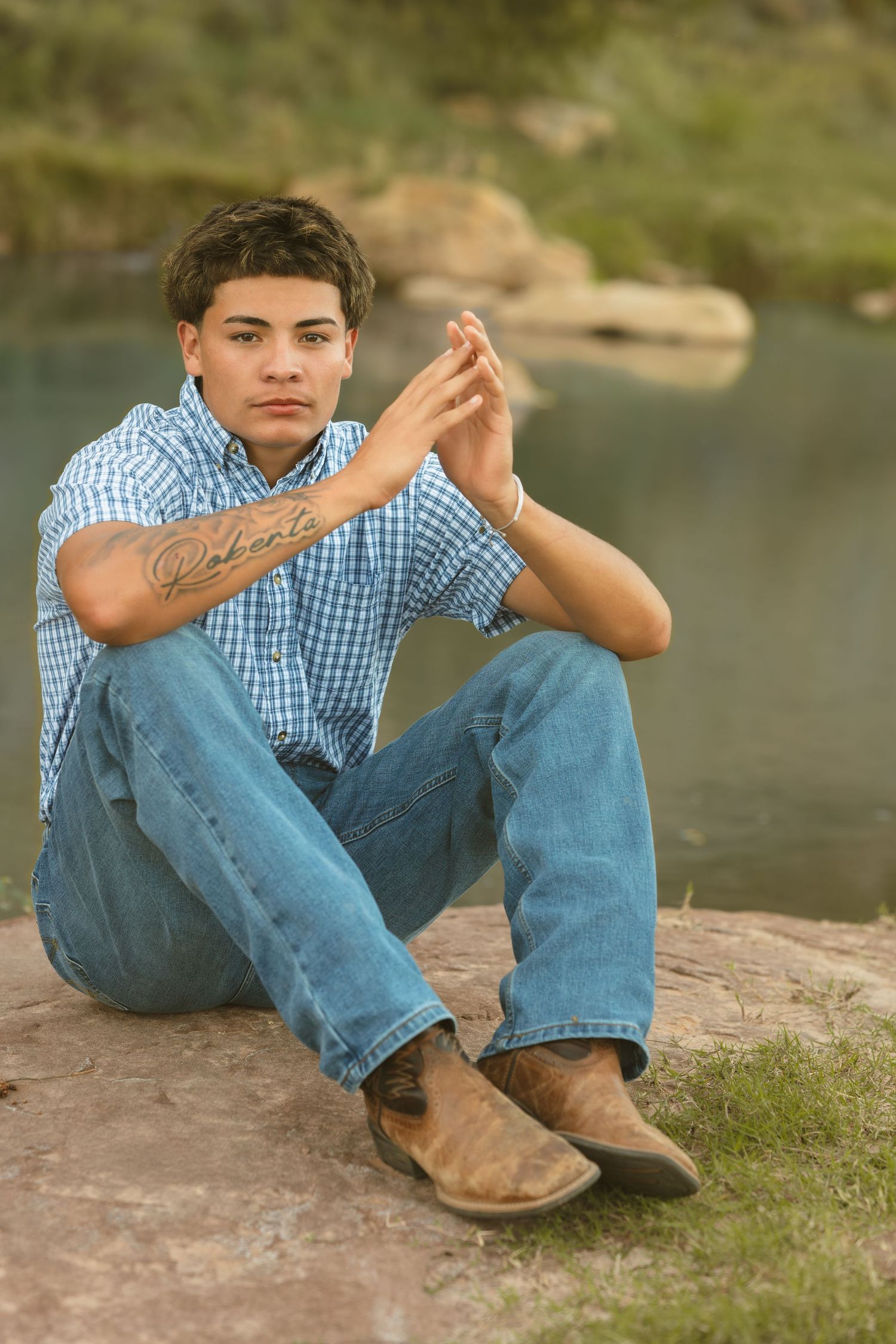Young man with forearm tattoo sitting on rocks near a river in a blue plaid shirt and jeans.