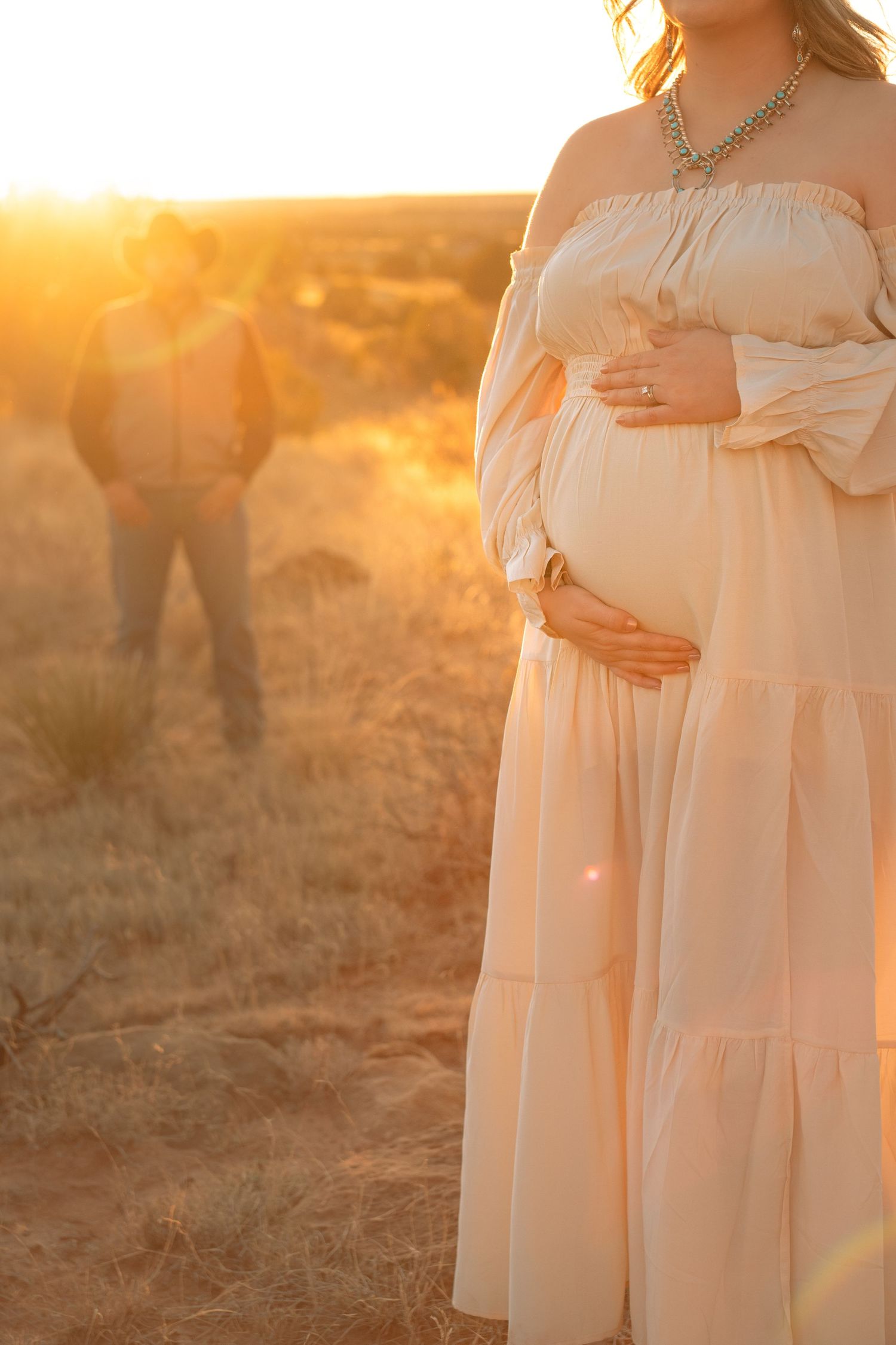 Pregnant woman in off-shoulder peach dress holding baby bump at golden sunset with man in background.