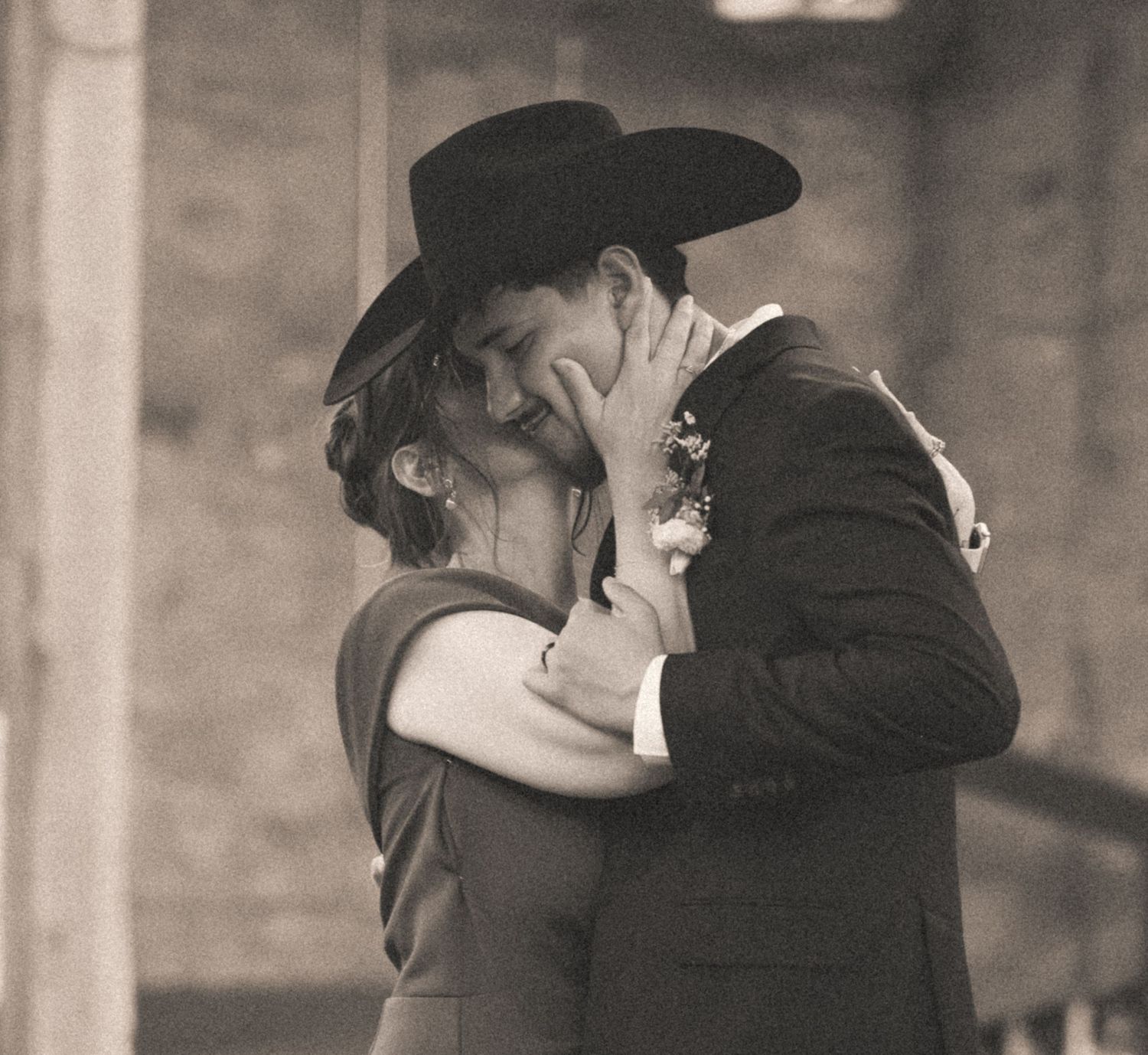 Groom in black cowboy hat tenderly kissing bride during romantic wedding ceremony in sepia tone.