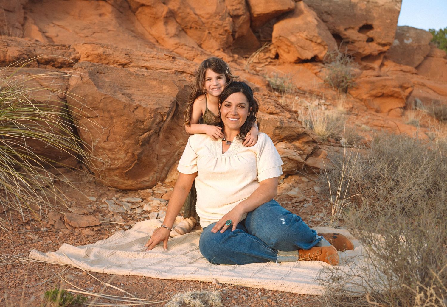 Woman and child sitting on blanket in front of red rock formations in desert landscape.