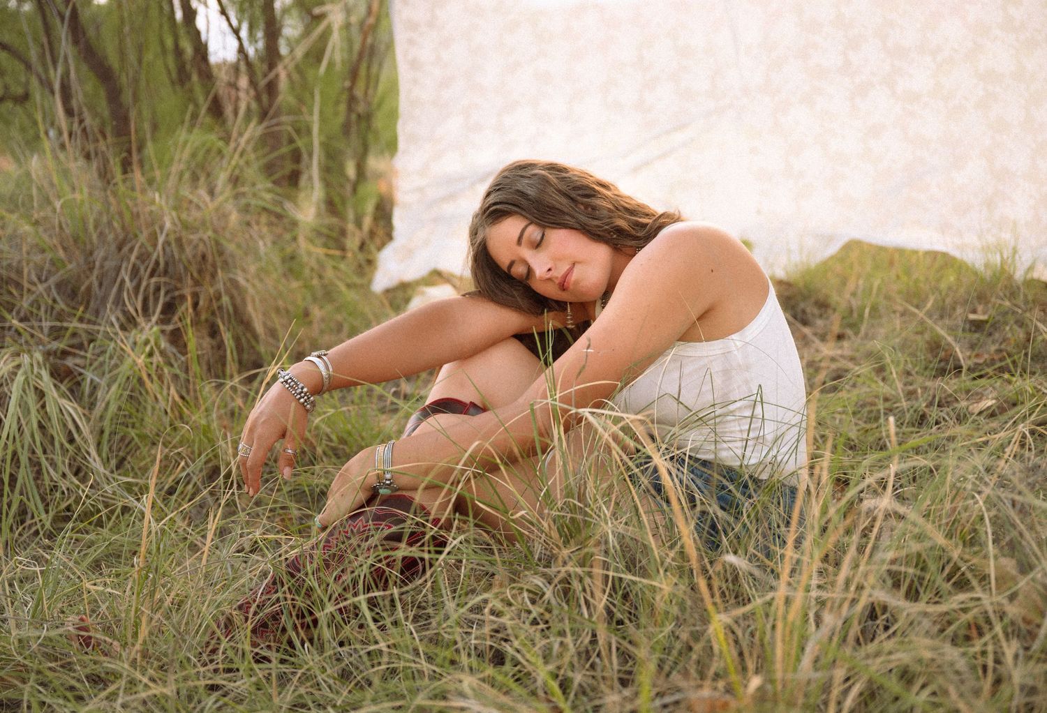 A young woman in a white tank top sits relaxed in tall grass outdoors, leaning on her knee in a serene natural setting.