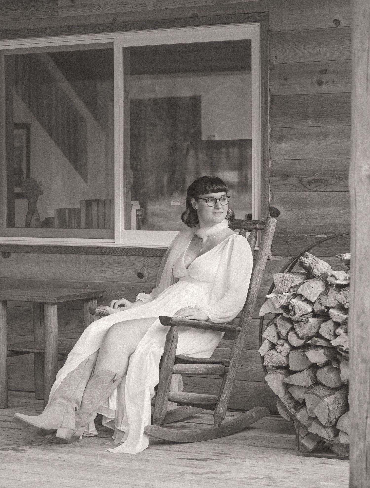Bride in white gown sitting in rustic rocking chair on log cabin porch in black and white.