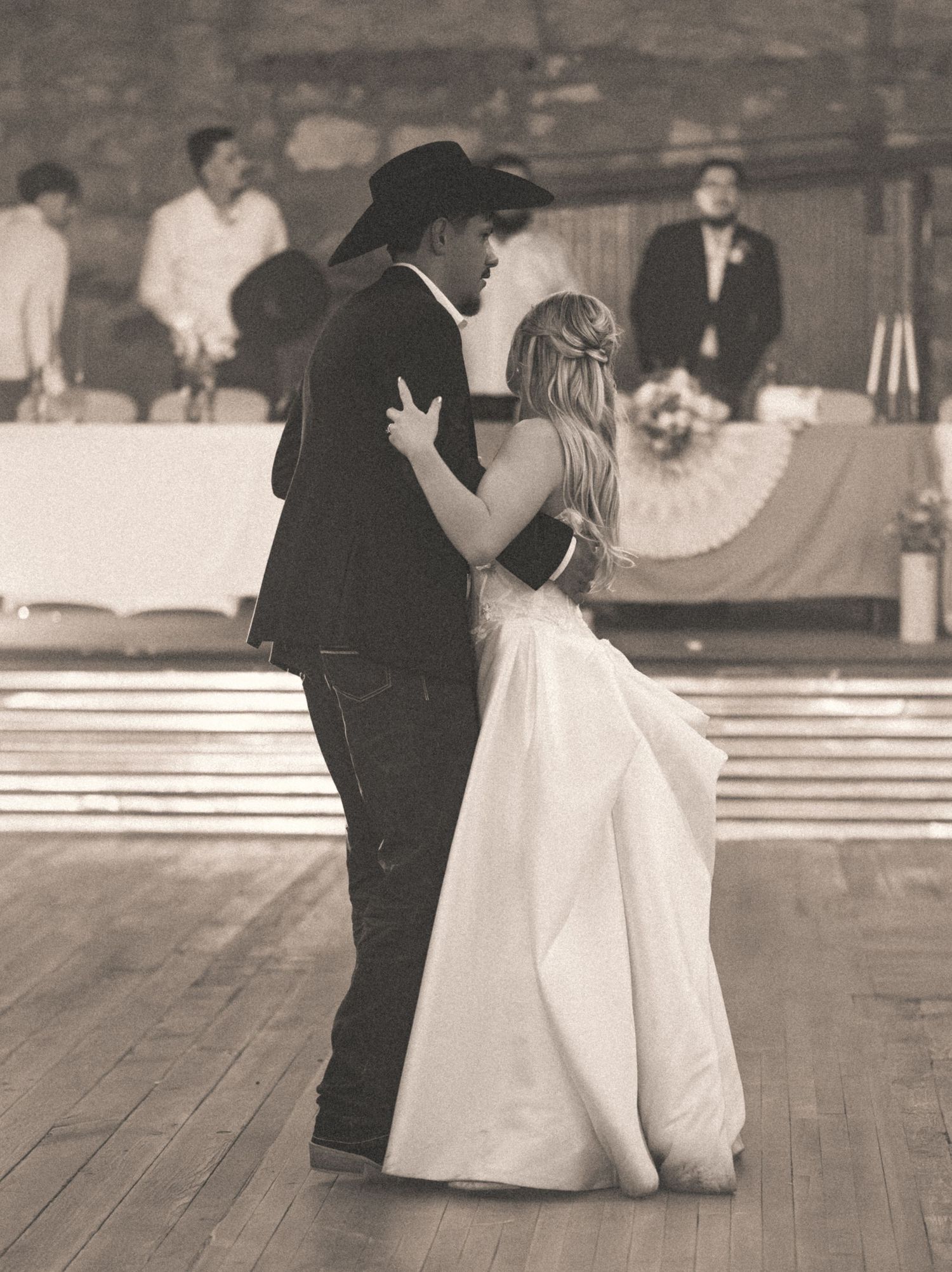 Bride and groom sharing first dance in cowboy hat at rustic wedding venue in sepia tone.