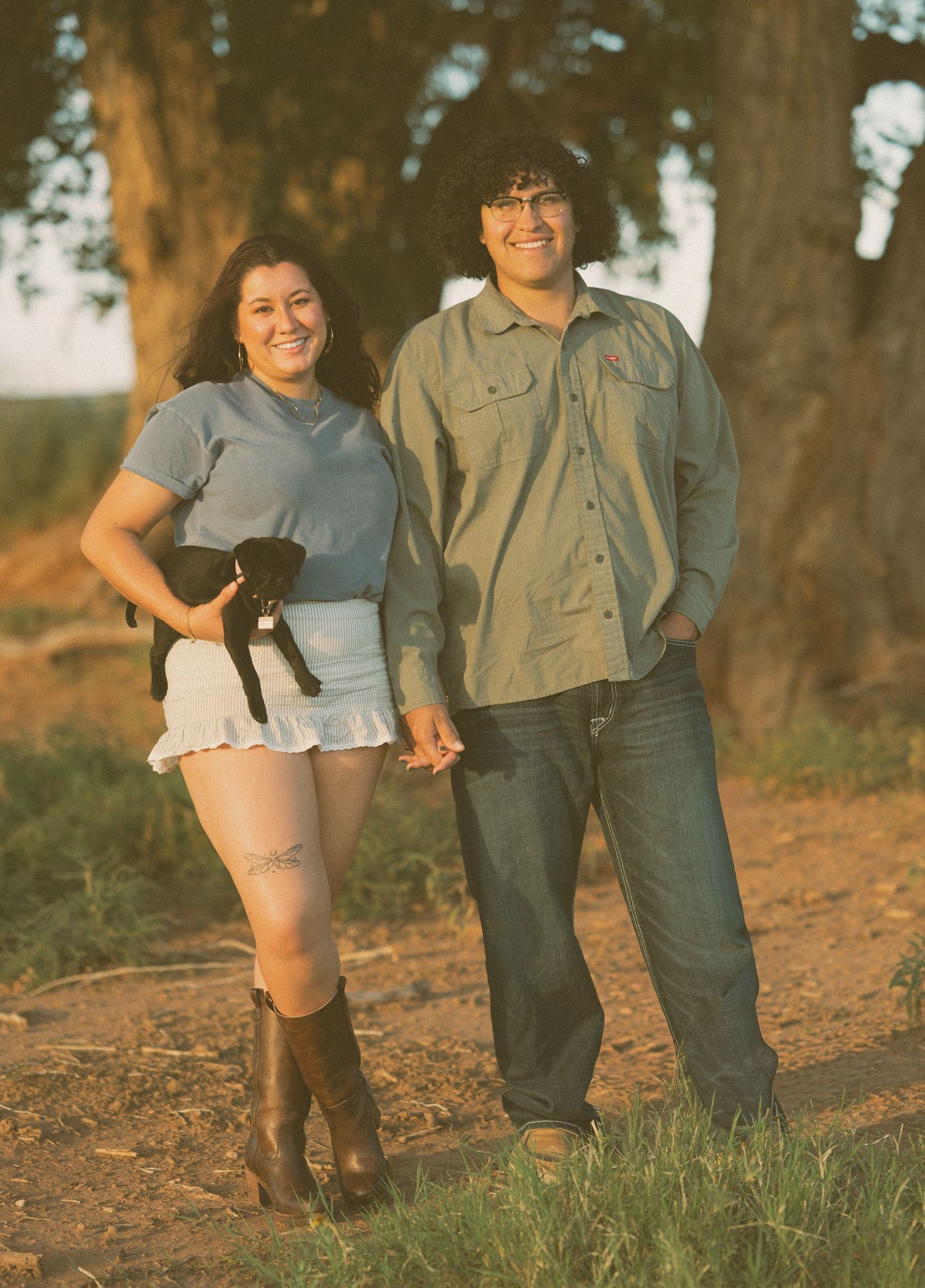 Smiling couple holding a black puppy while standing on a dirt path at golden hour.
