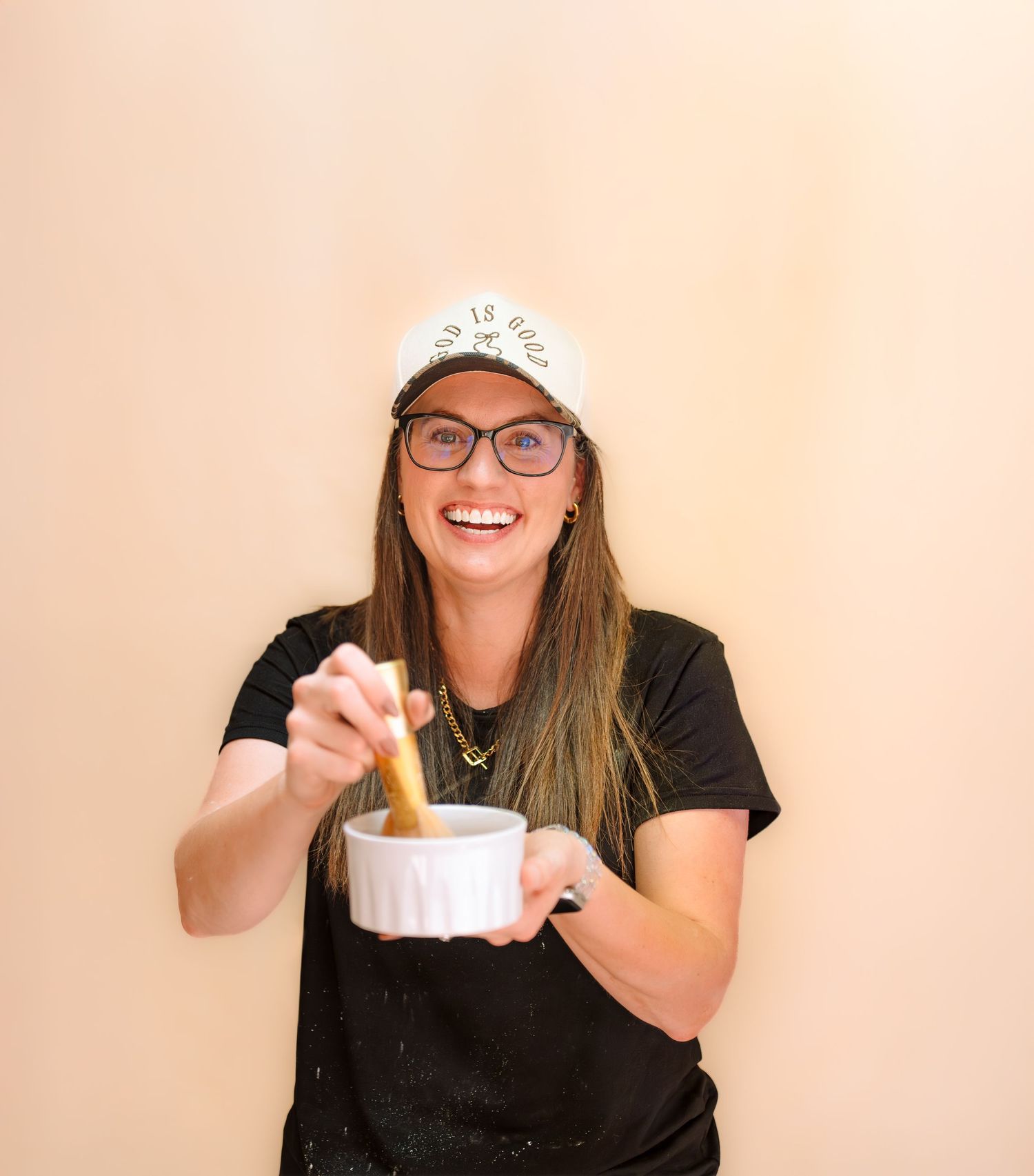 Smiling woman wearing glasses and a white cap uses a pestle in a white mortar bowl against a beige background.