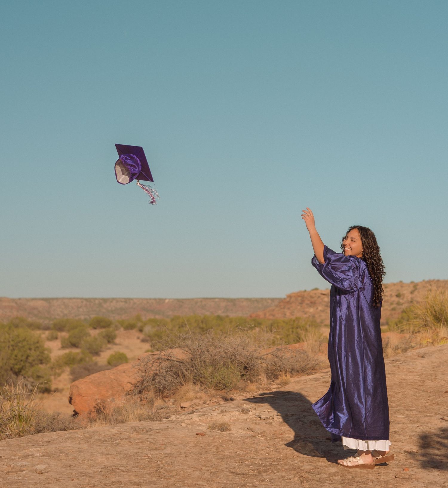 Graduate in purple gown tosses cap into air outdoors in desert landscape under clear blue sky.