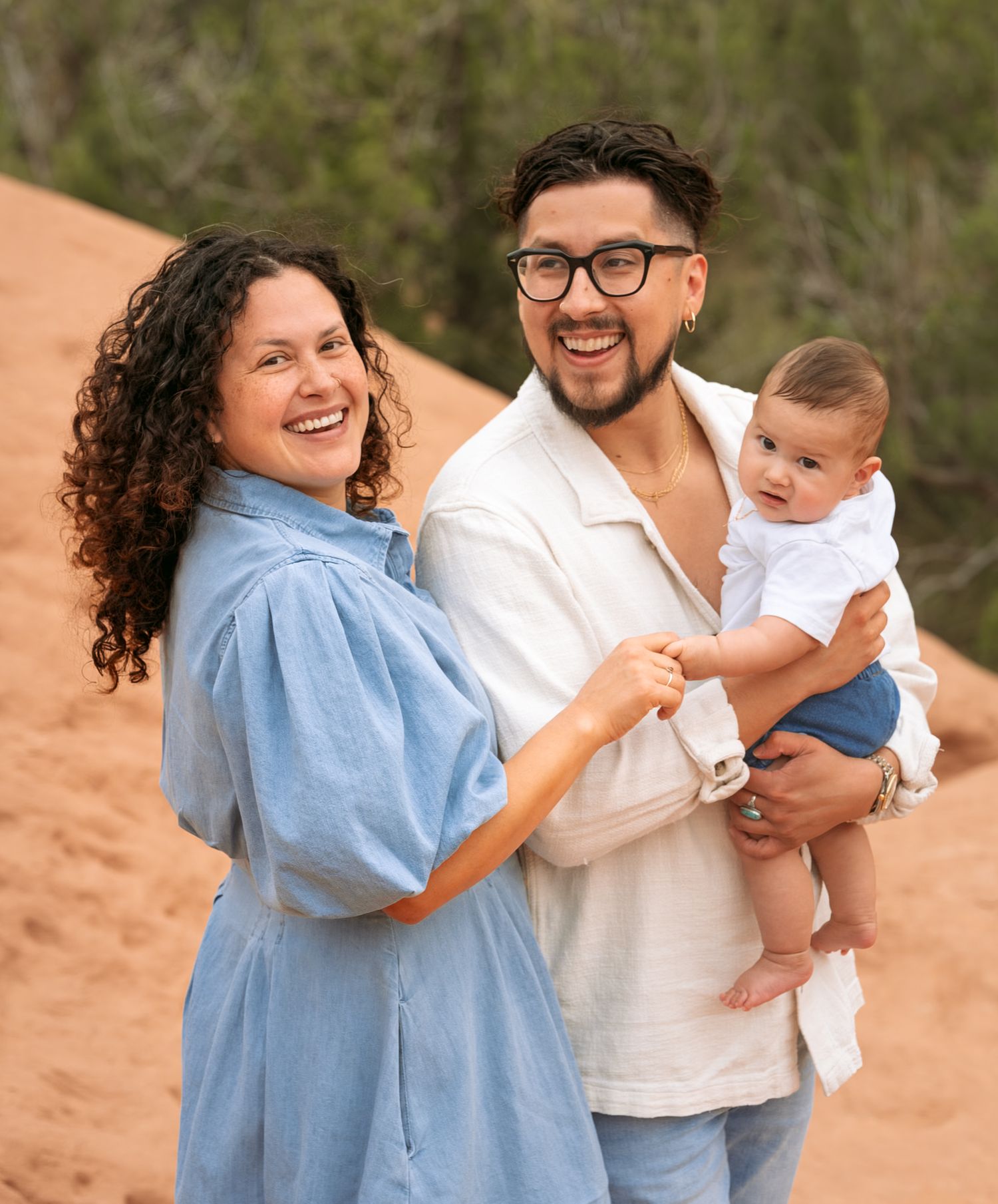 Smiling couple holding infant baby outdoors on sandy dunes in casual blue outfits.