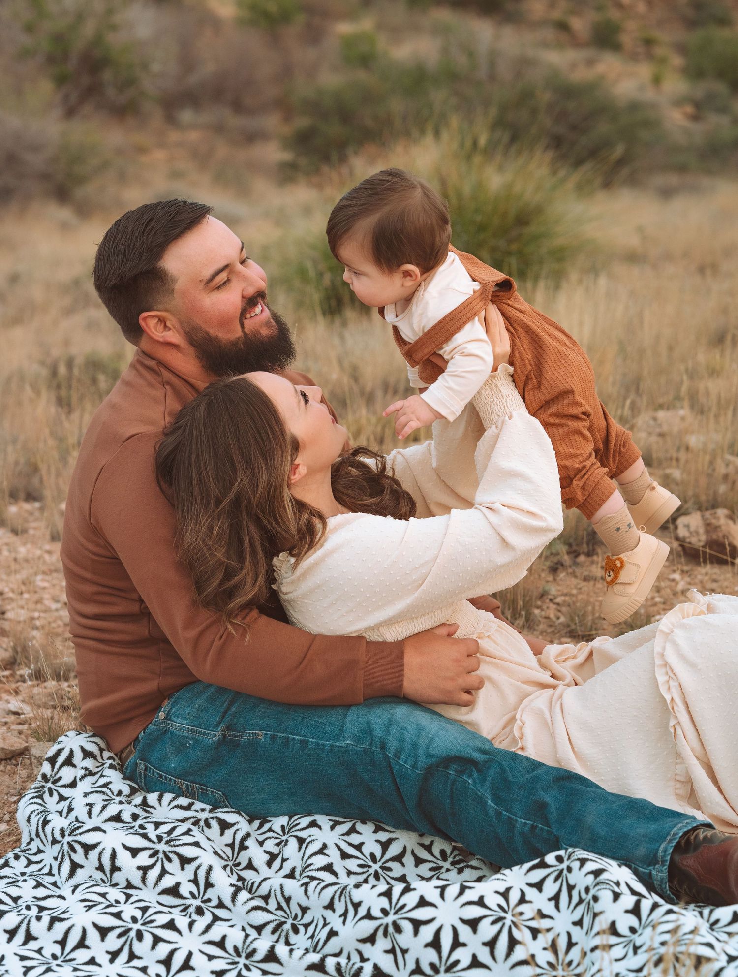 Young family sitting on floral blanket in desert field with parents lifting laughing baby between them.