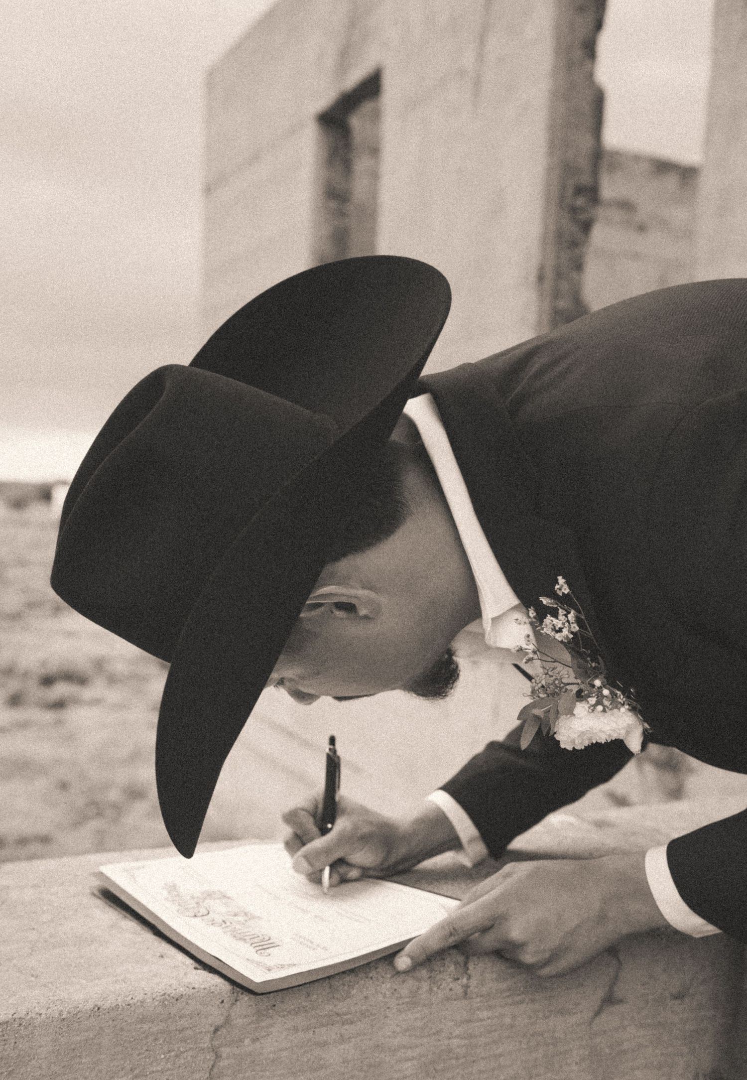 Groom in black cowboy hat and suit signing wedding certificate at outdoor rustic ceremony.