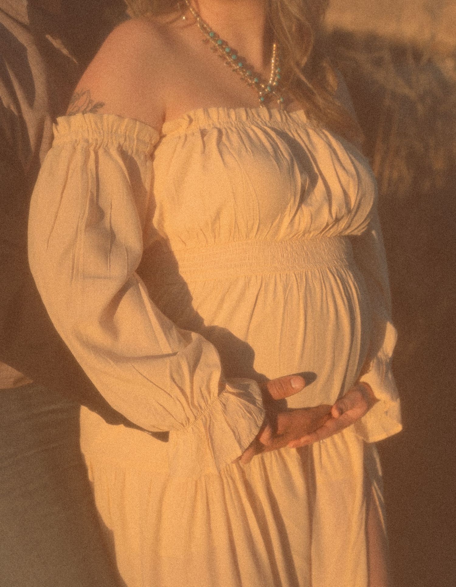 Close-up of pregnant woman in off-shoulder cream dress cradling baby bump with layered necklace.