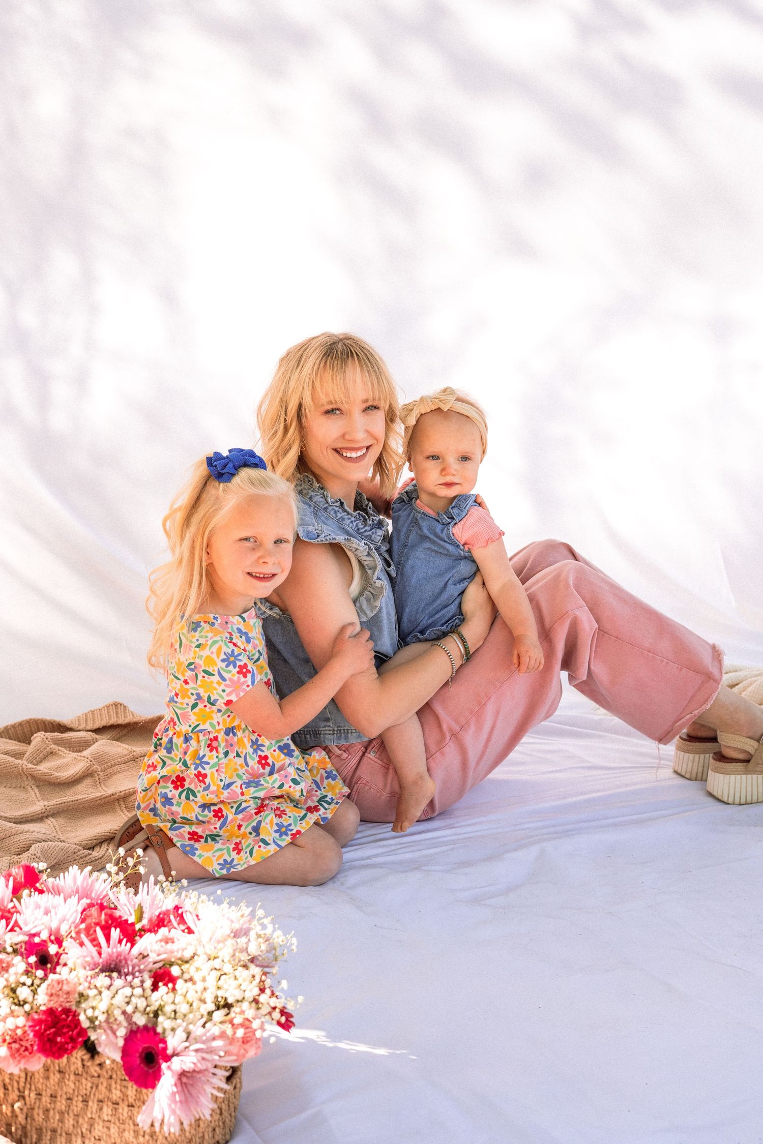 Smiling blonde mother sits with two young daughters on white backdrop, surrounded by pink flowers in a wicker basket.