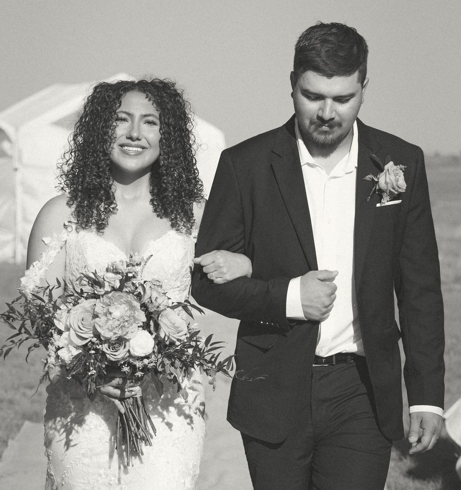 Smiling bride with curly hair and groom walk arm in arm in black and white.