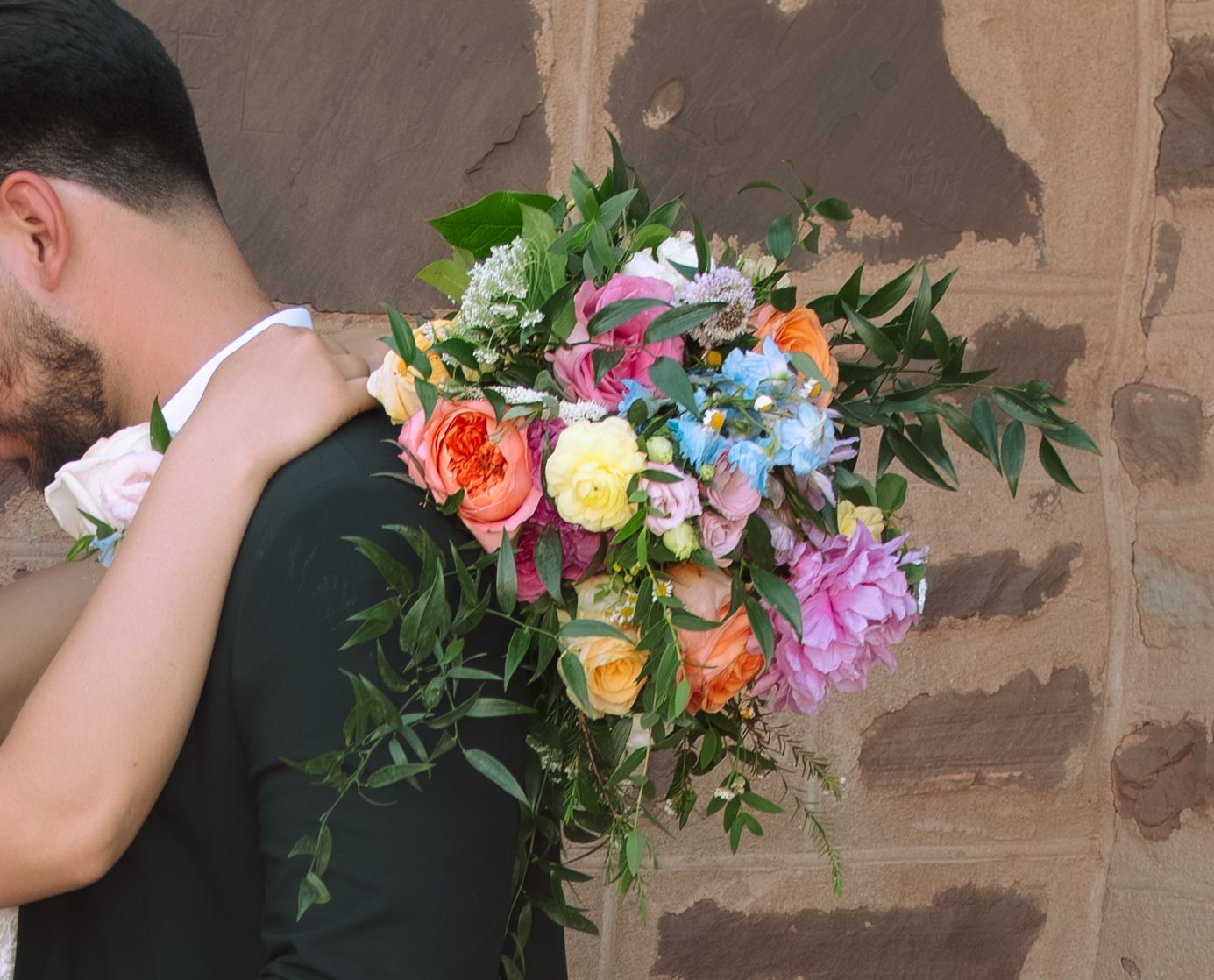 Groom holds a vibrant colorful bridal bouquet with roses against a stone wall.