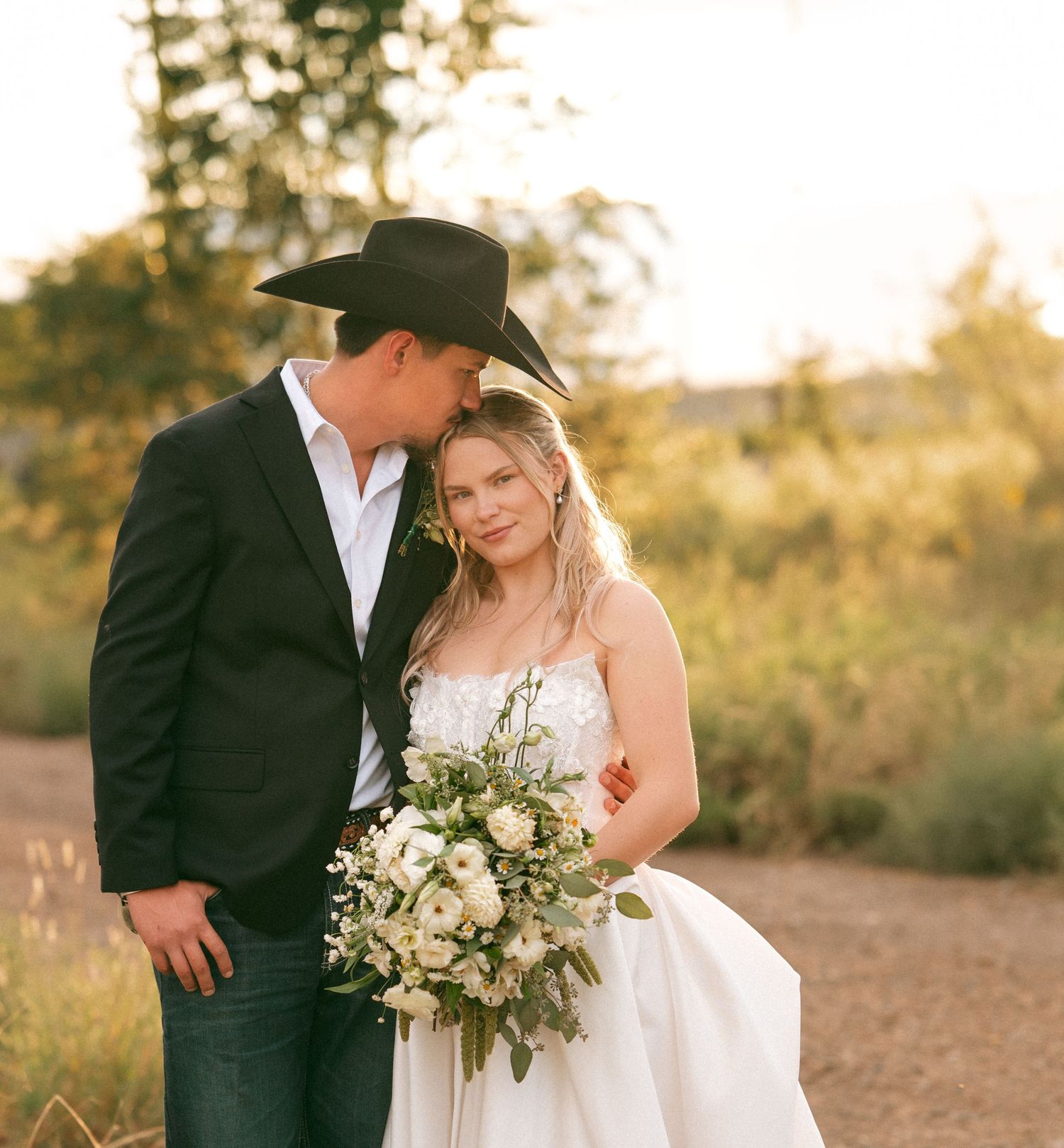 Bride and groom in cowboy hat sharing intimate moment in sun-drenched outdoor field setting.