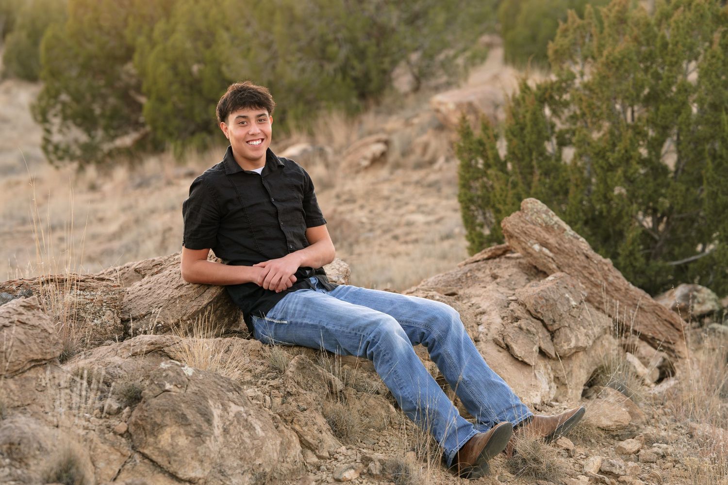 Young man in black shirt and jeans sitting relaxed on rocks in a desert landscape with shrubs.