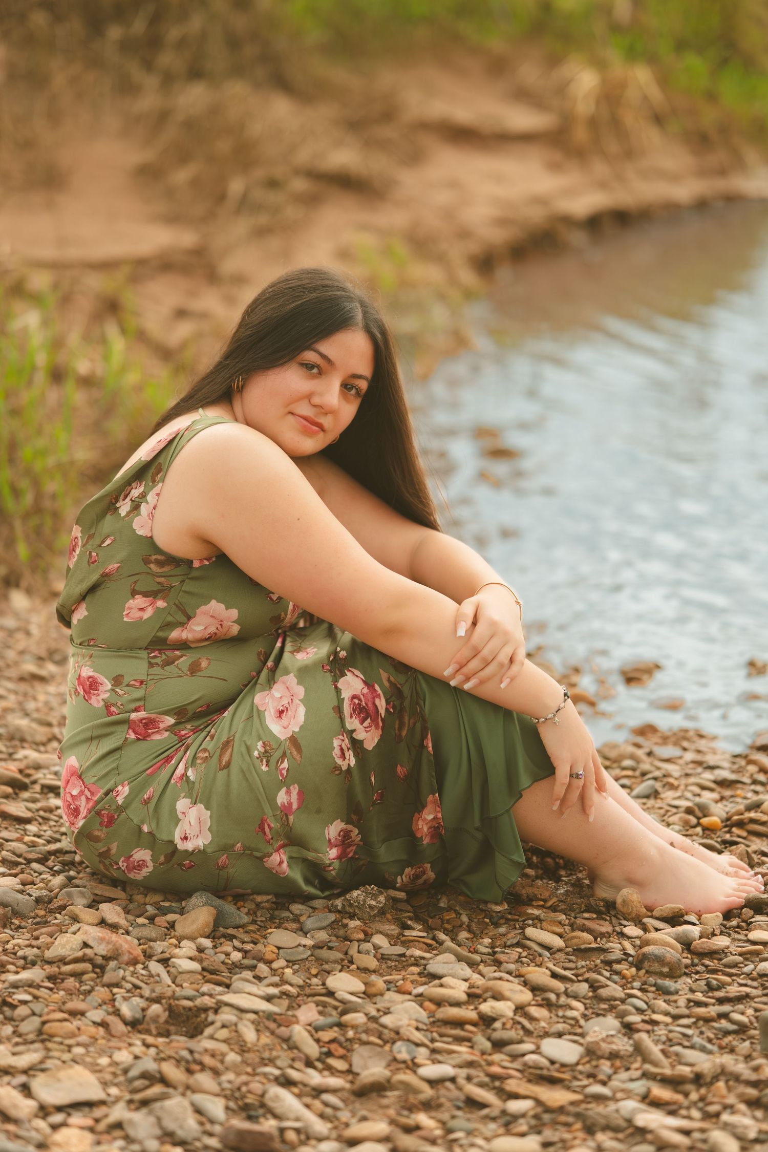 Young woman in floral green dress sits barefoot on rocky riverbank smiling at camera outdoors.