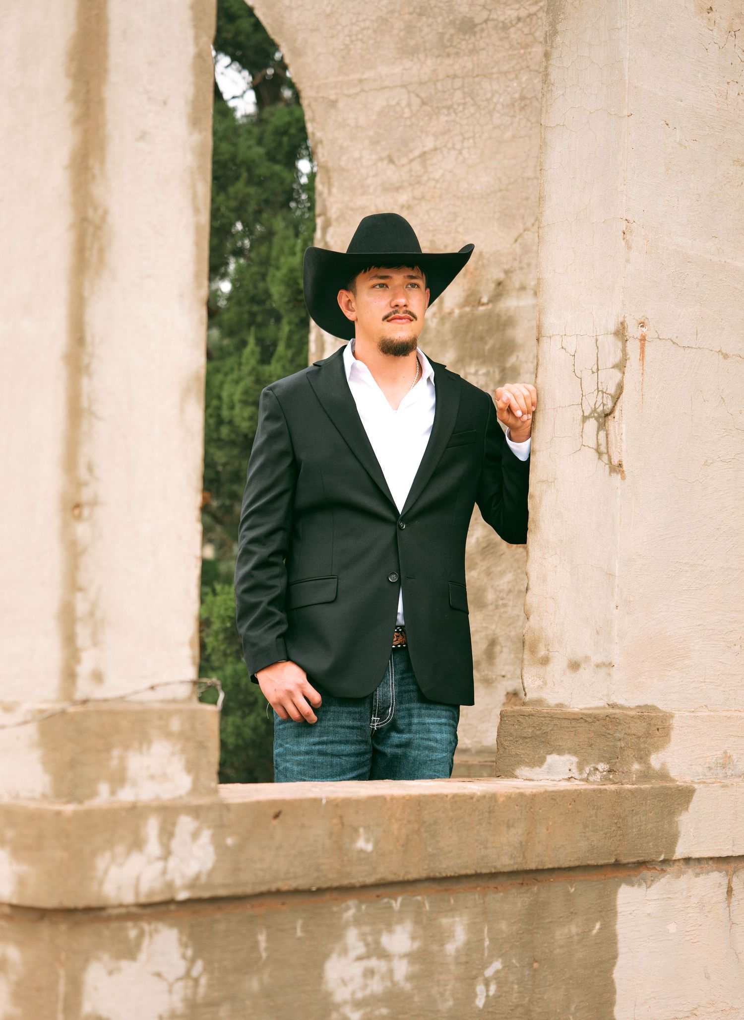 Young man in black cowboy hat and blazer with jeans posing between stone archways outdoors.