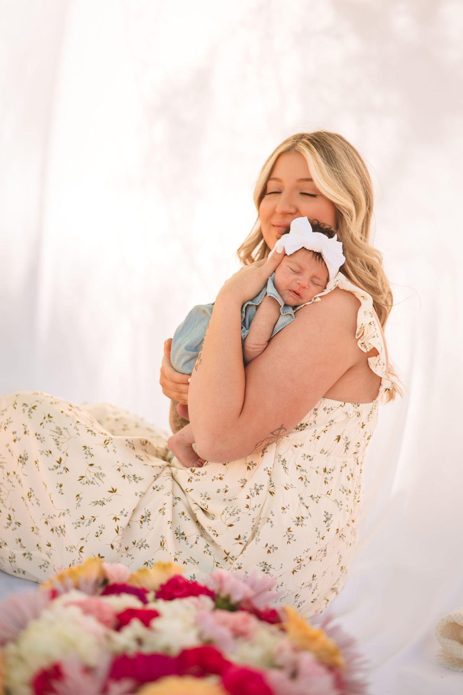 A mother in a floral dress tenderly holds her sleeping newborn with a white bow against a soft white background.
