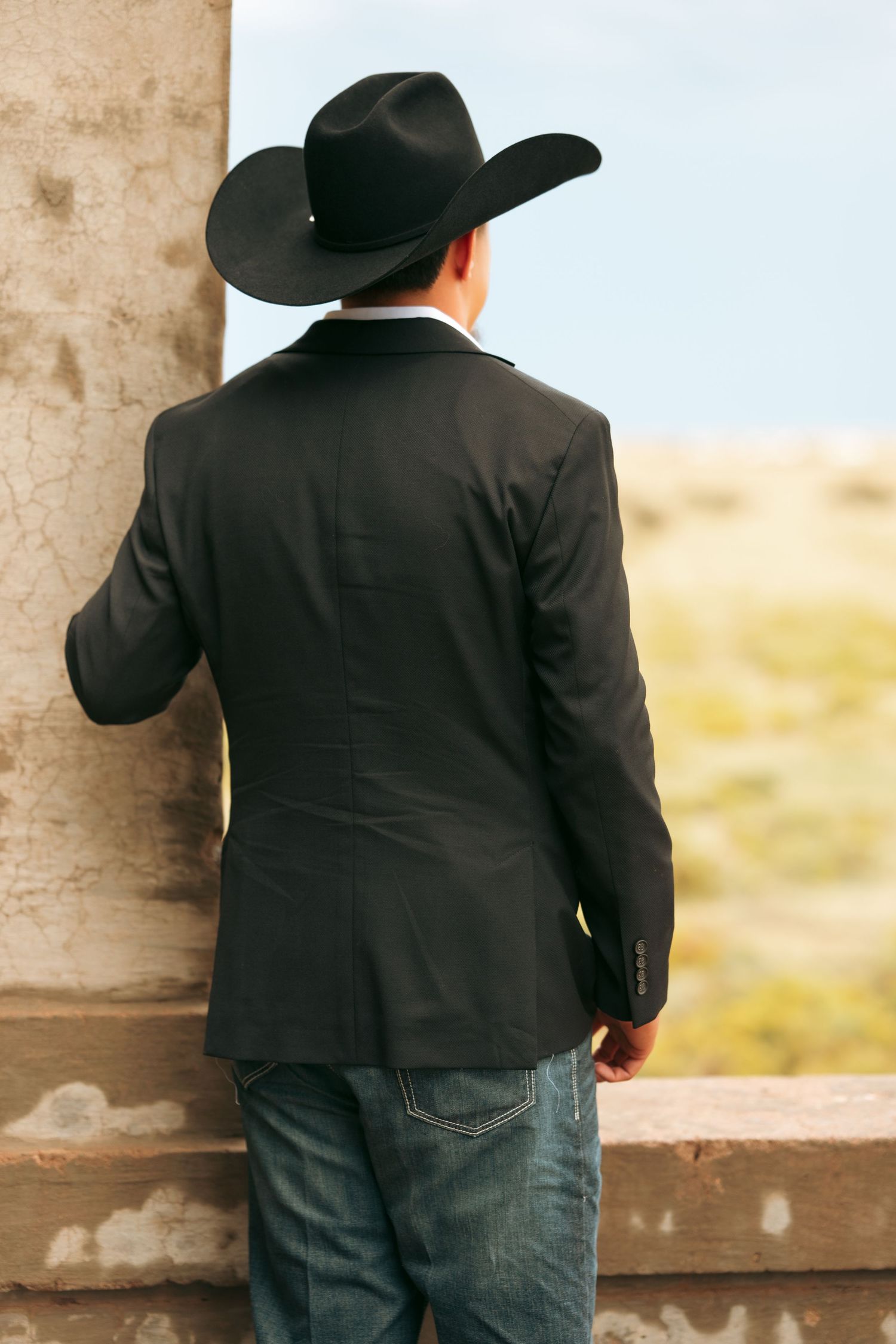 Young man in black cowboy hat and blazer with jeans posing between stone archways outdoors.
