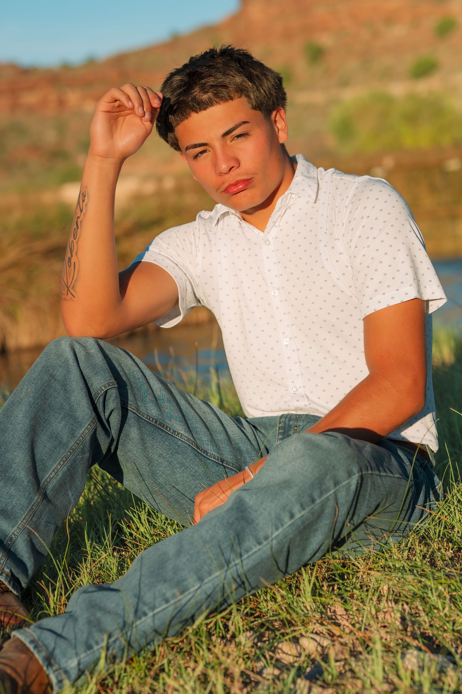 Young man with tattoo sitting in grass at golden hour near water, wearing white polo and jeans.
