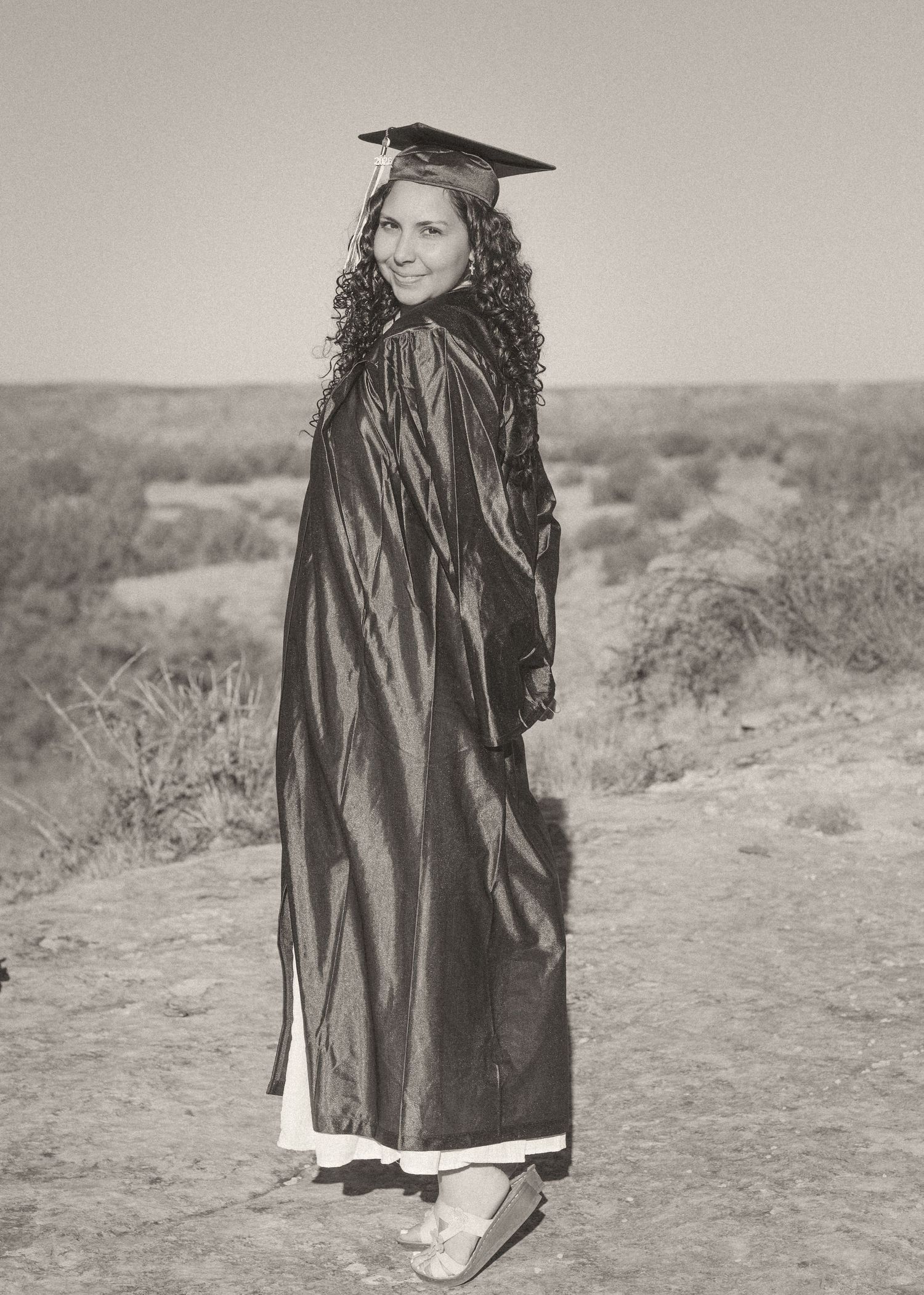 A young woman in black graduation gown and cap poses outdoors in a desert landscape, smiling over her shoulder.