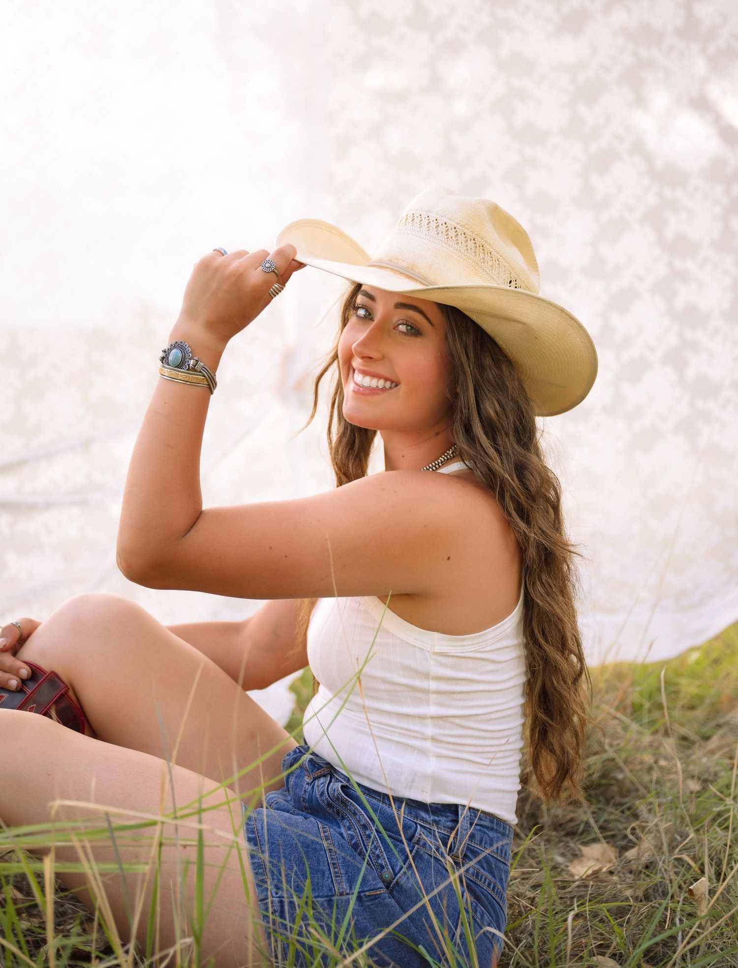 Smiling young woman in white tank top and cowboy hat sitting in grass, wearing denim shorts and turquoise bracelet.