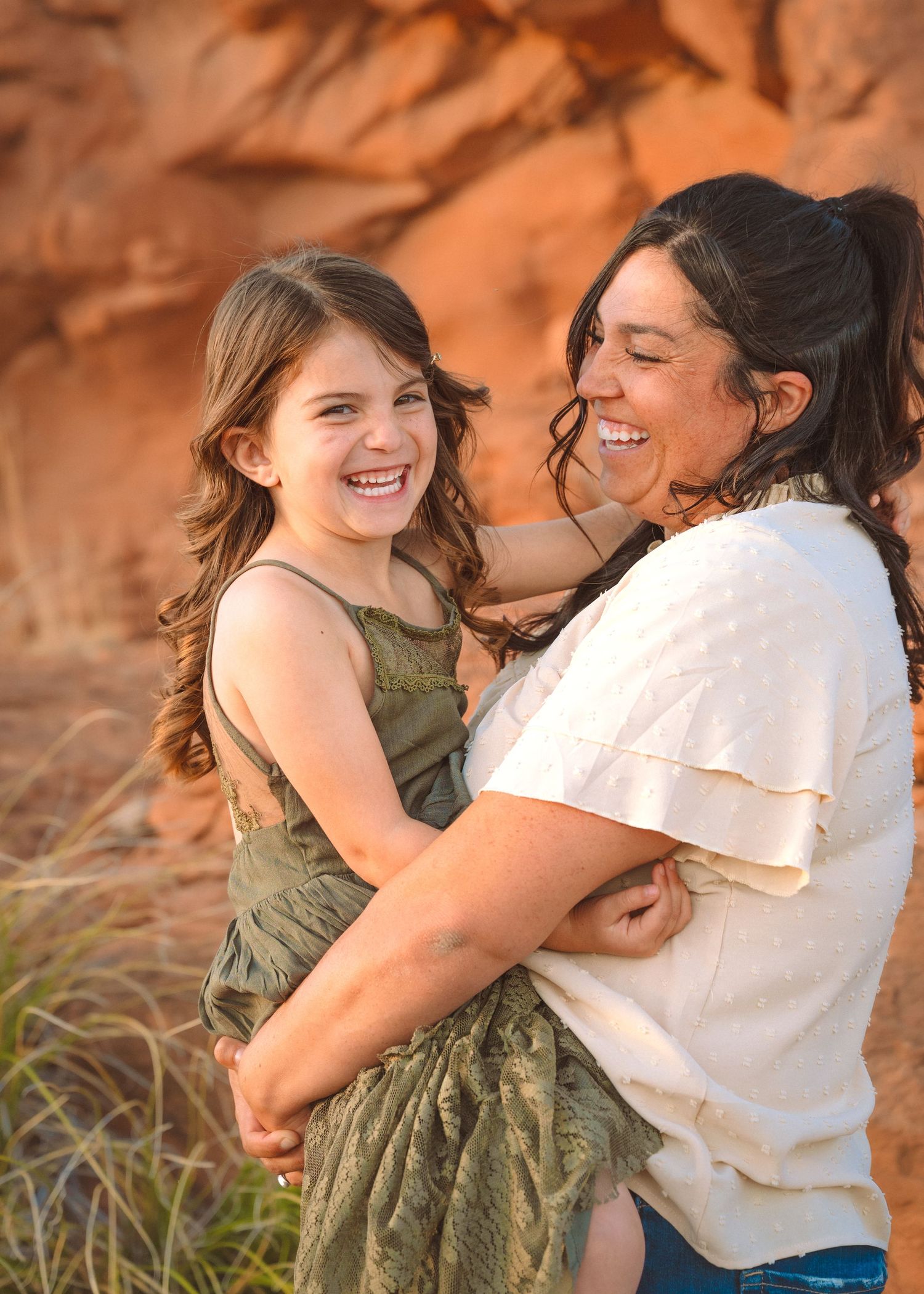 A joyful mother holds her laughing young daughter outdoors near red rock formations in warm golden light.