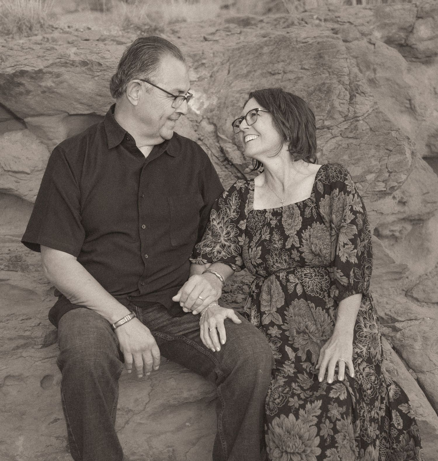 Couple sitting on rocks holding hands and gazing at each other in a sepia-toned outdoor portrait.