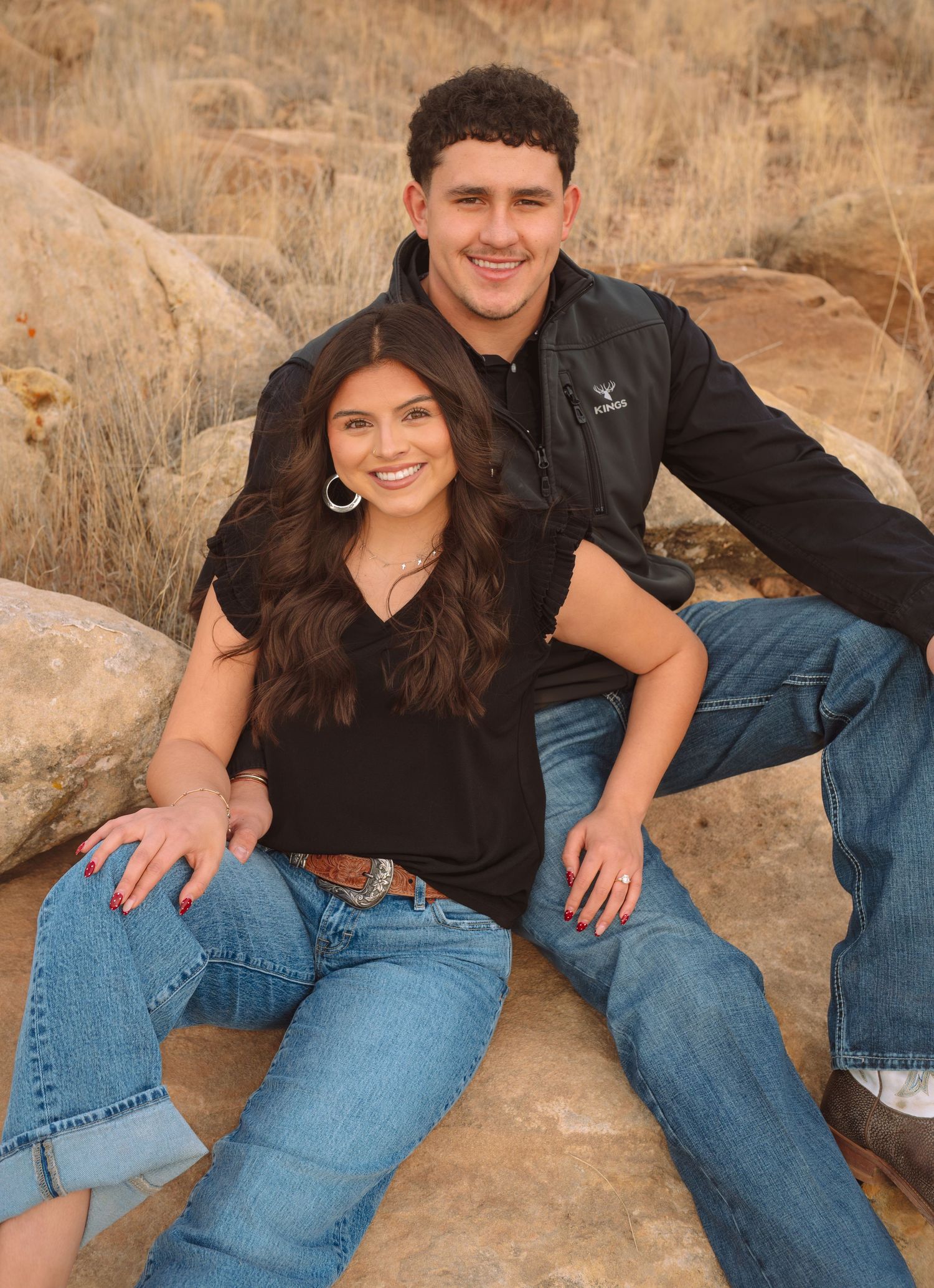 Smiling teenage couple posing on desert rocks, girl in black top and boy in dark vest and jeans.