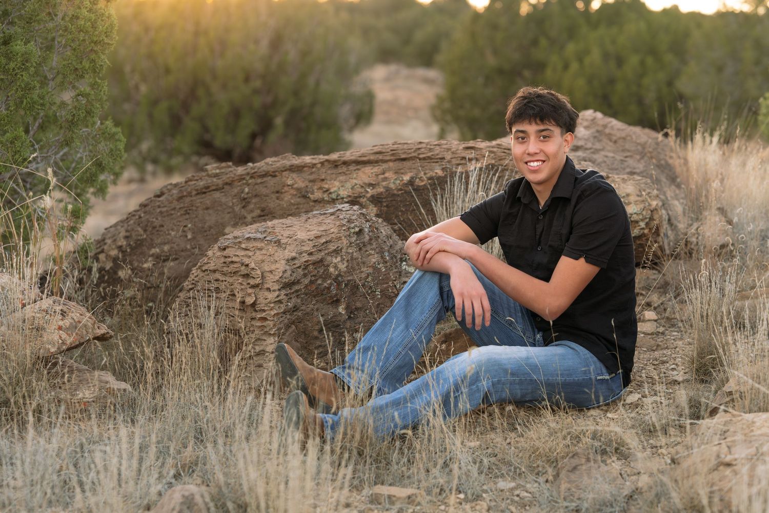 Smiling teenage boy sits on desert rocks surrounded by dry grass and juniper trees at sunset.