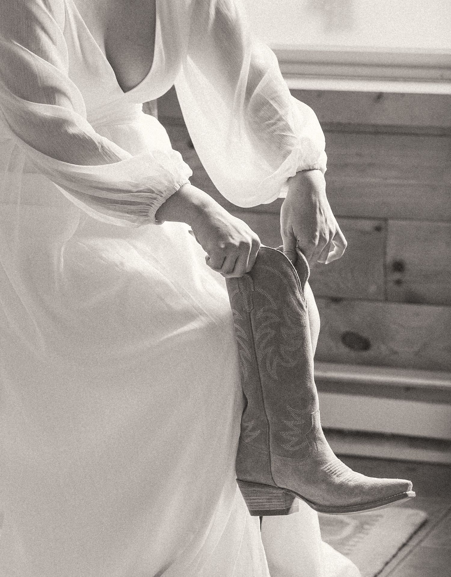 Bride in white wedding dress pulling on embroidered cowboy boots in black and white photo.