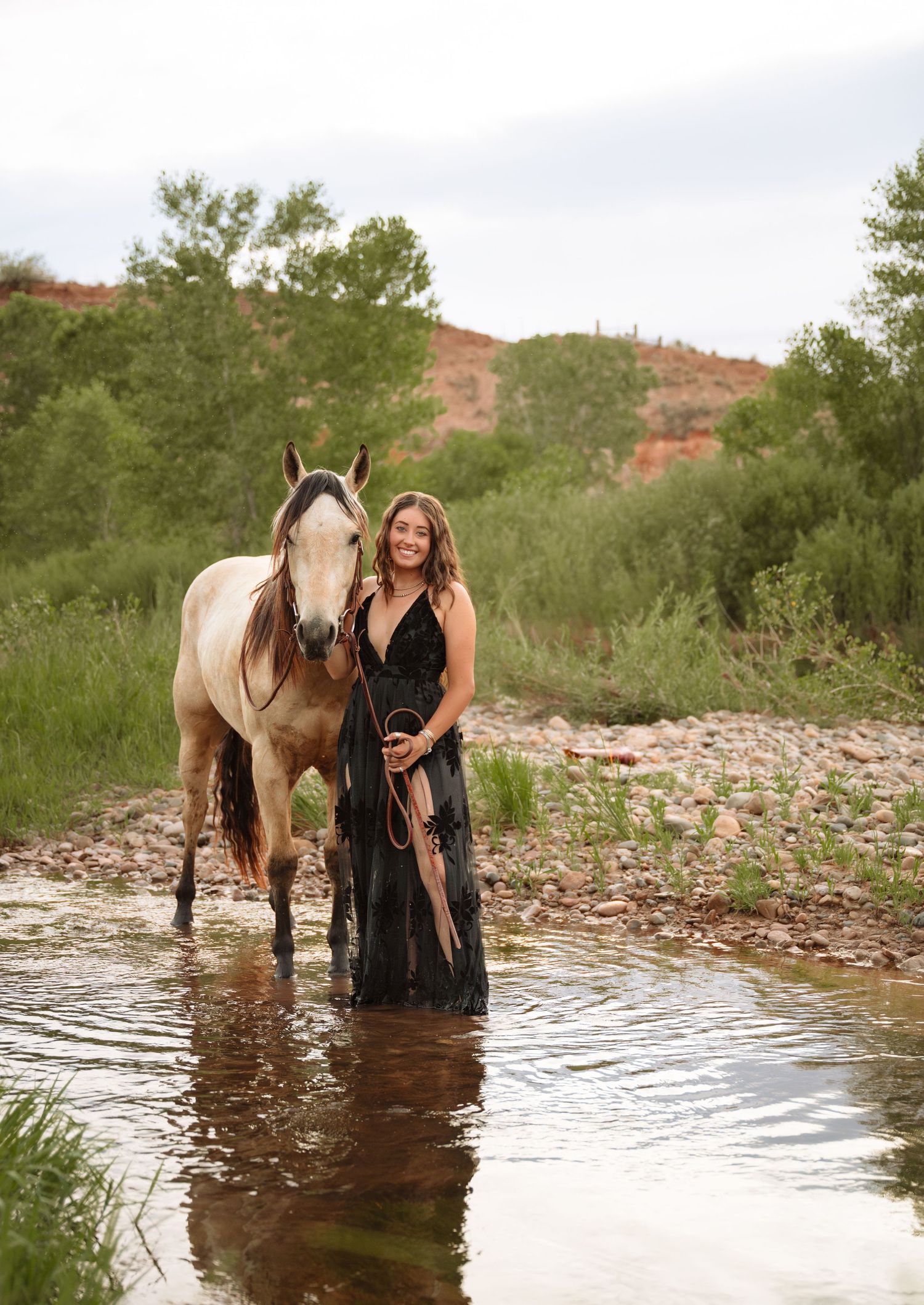 Young woman in black gown stands with buckskin horse in a shallow creek surrounded by lush greenery and red rock hills.
