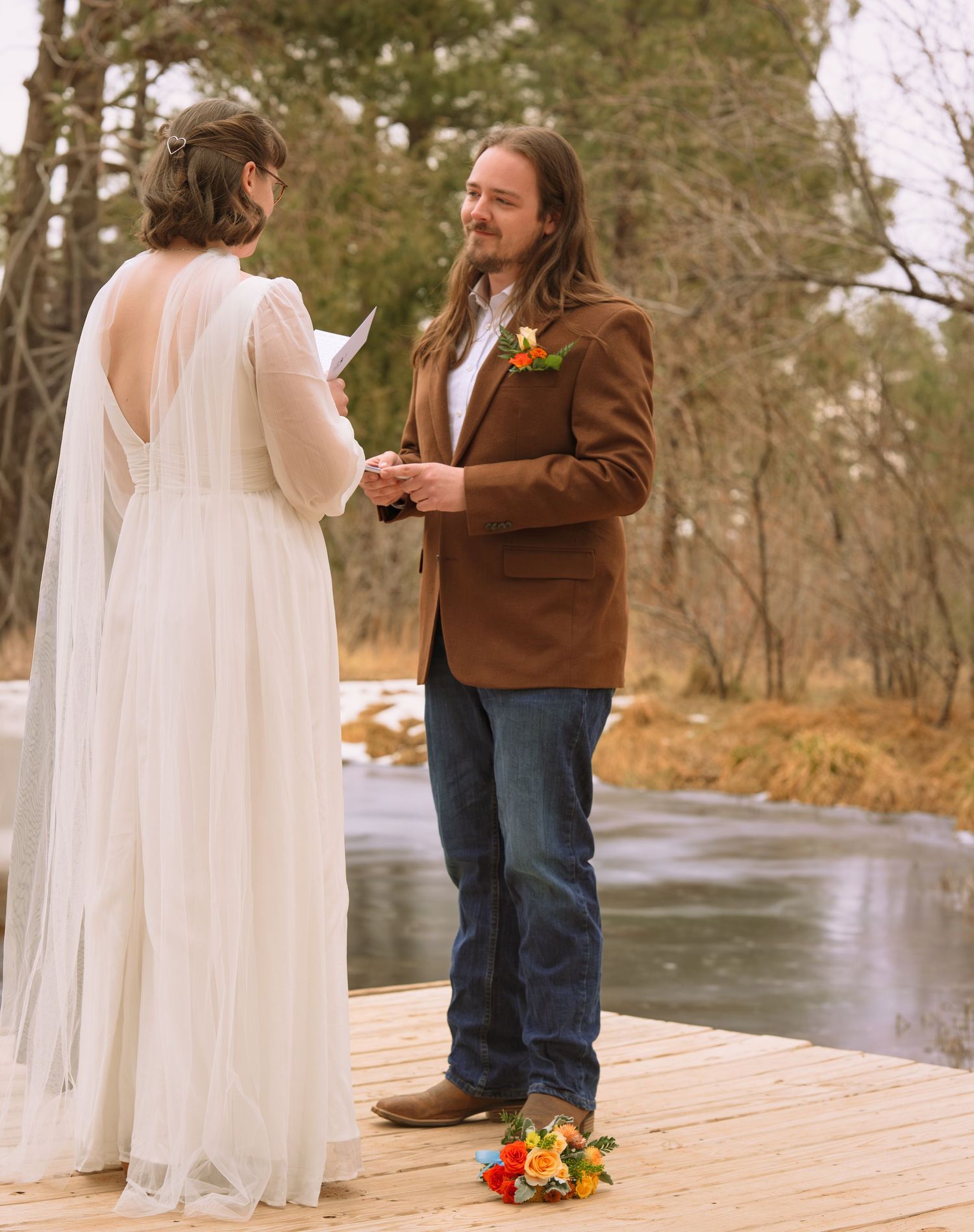 Bride reading vows to groom in brown blazer on wooden dock with snowy river backdrop. Elopement in Torreon, New Mexico,