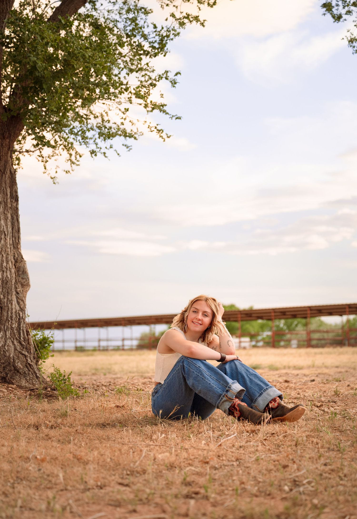 A smiling woman sits relaxed under a large tree in a rural field, wearing jeans and a beige top on a sunny day.