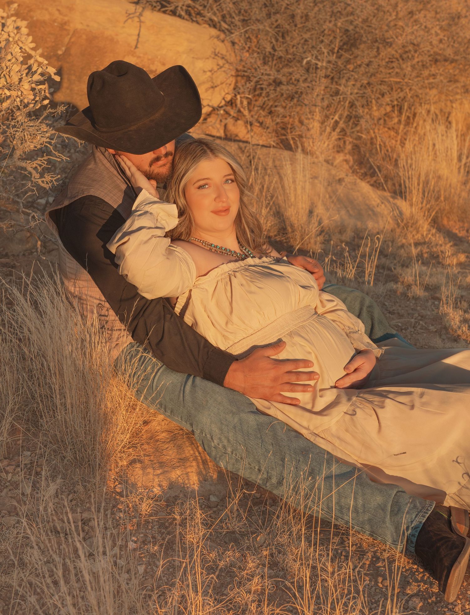 Pregnant woman reclining in mans arms in rustic desert setting, maternity photo at golden hour.