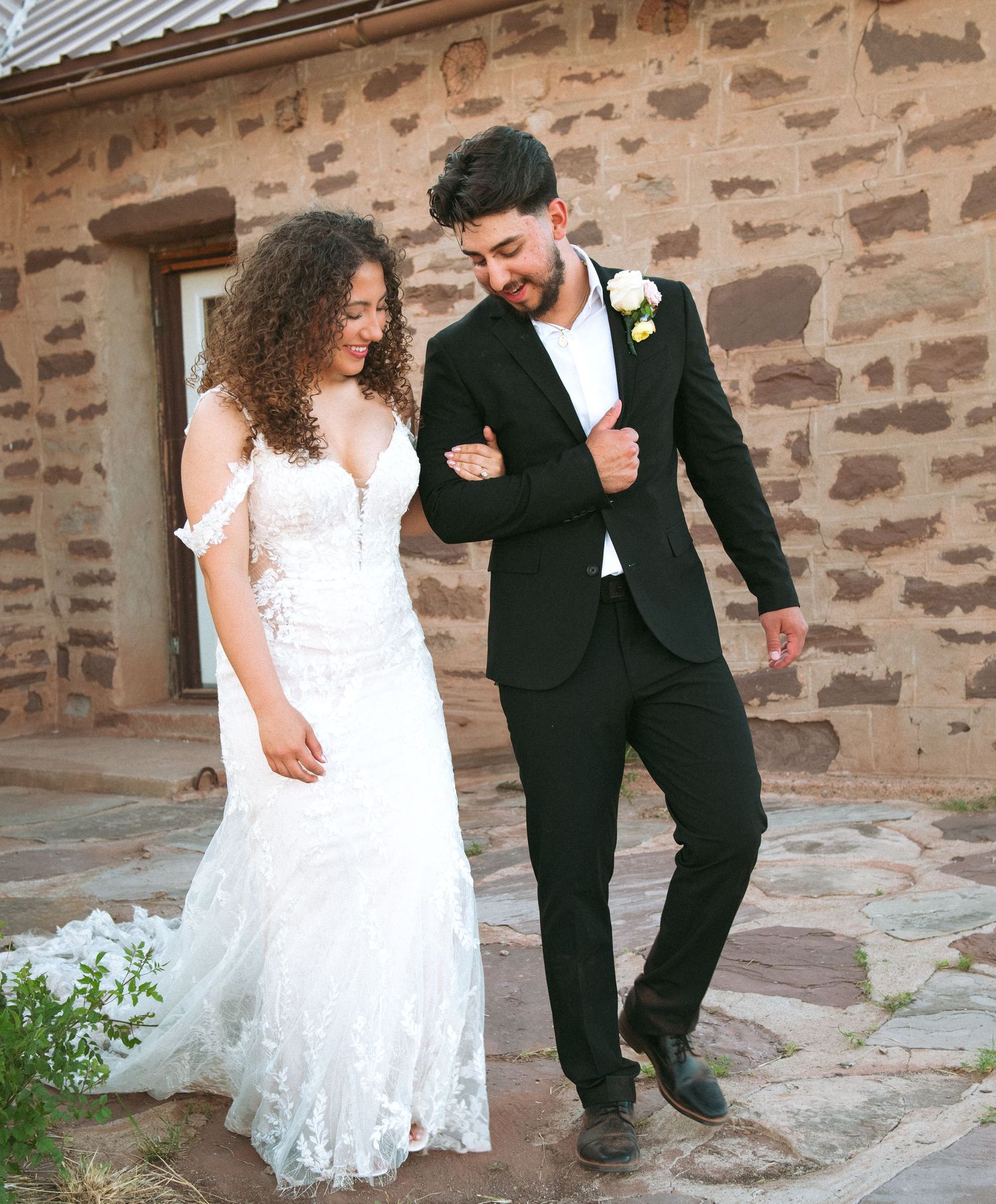 Smiling bride and groom in black suit walk together outside a stone building.