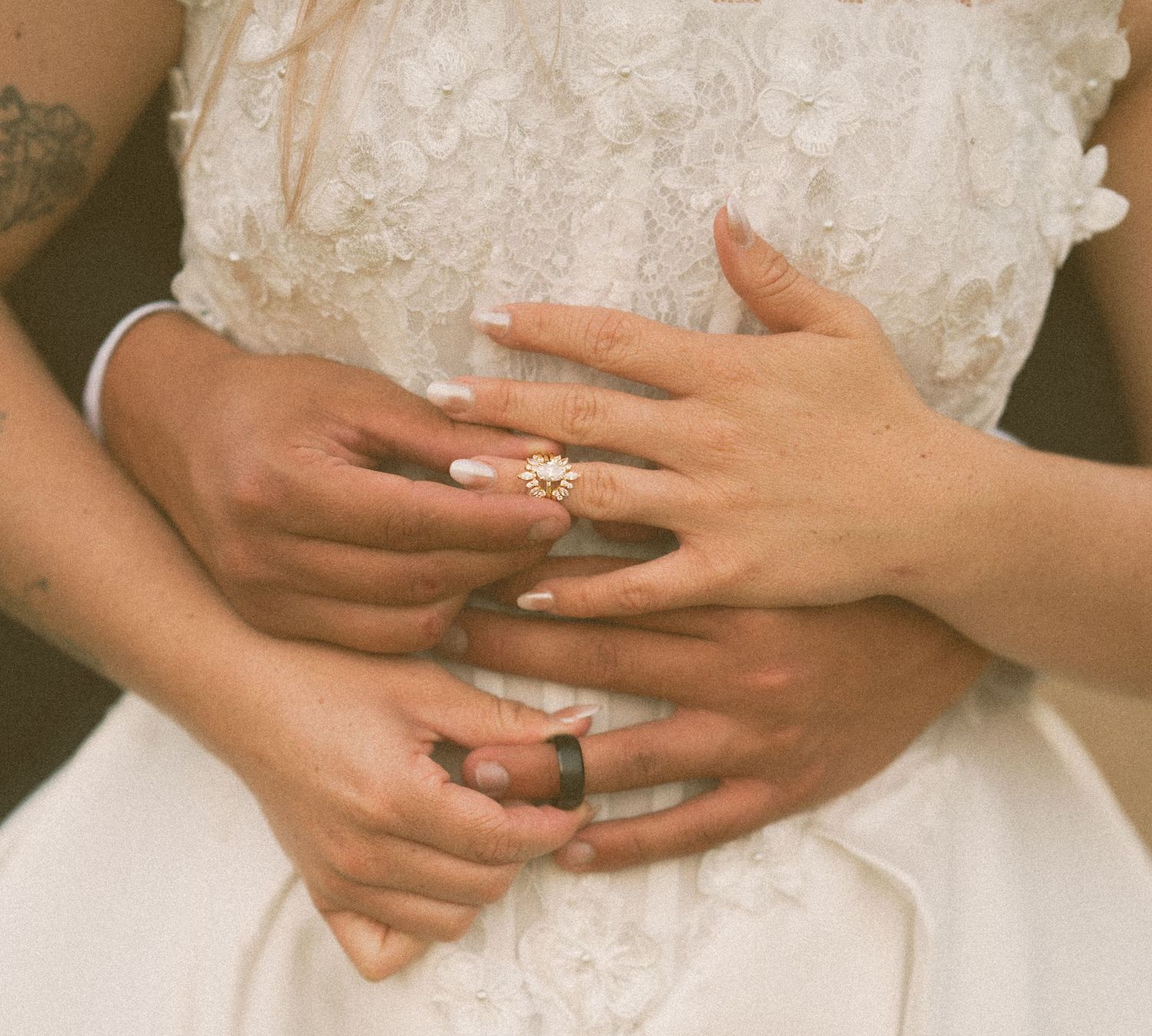 Close-up of couple\u2019s hands with floral engagement ring and wedding bands on bride\u2019s white gown.