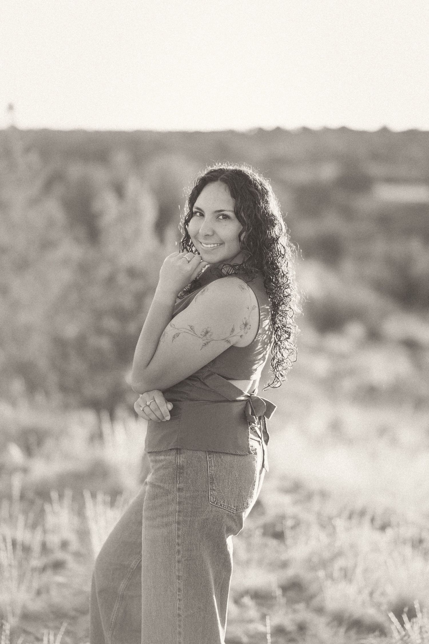 Black and white portrait of a smiling woman with curly hair posing outdoors in a field.