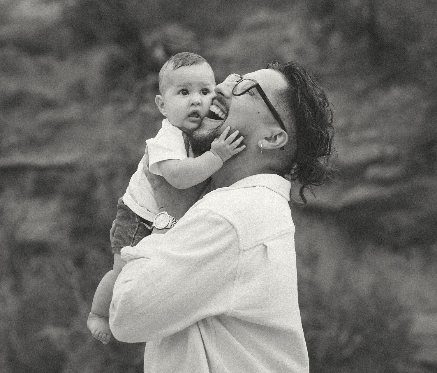A laughing man with sunglasses holds a curious baby outdoors in a black and white family portrait.